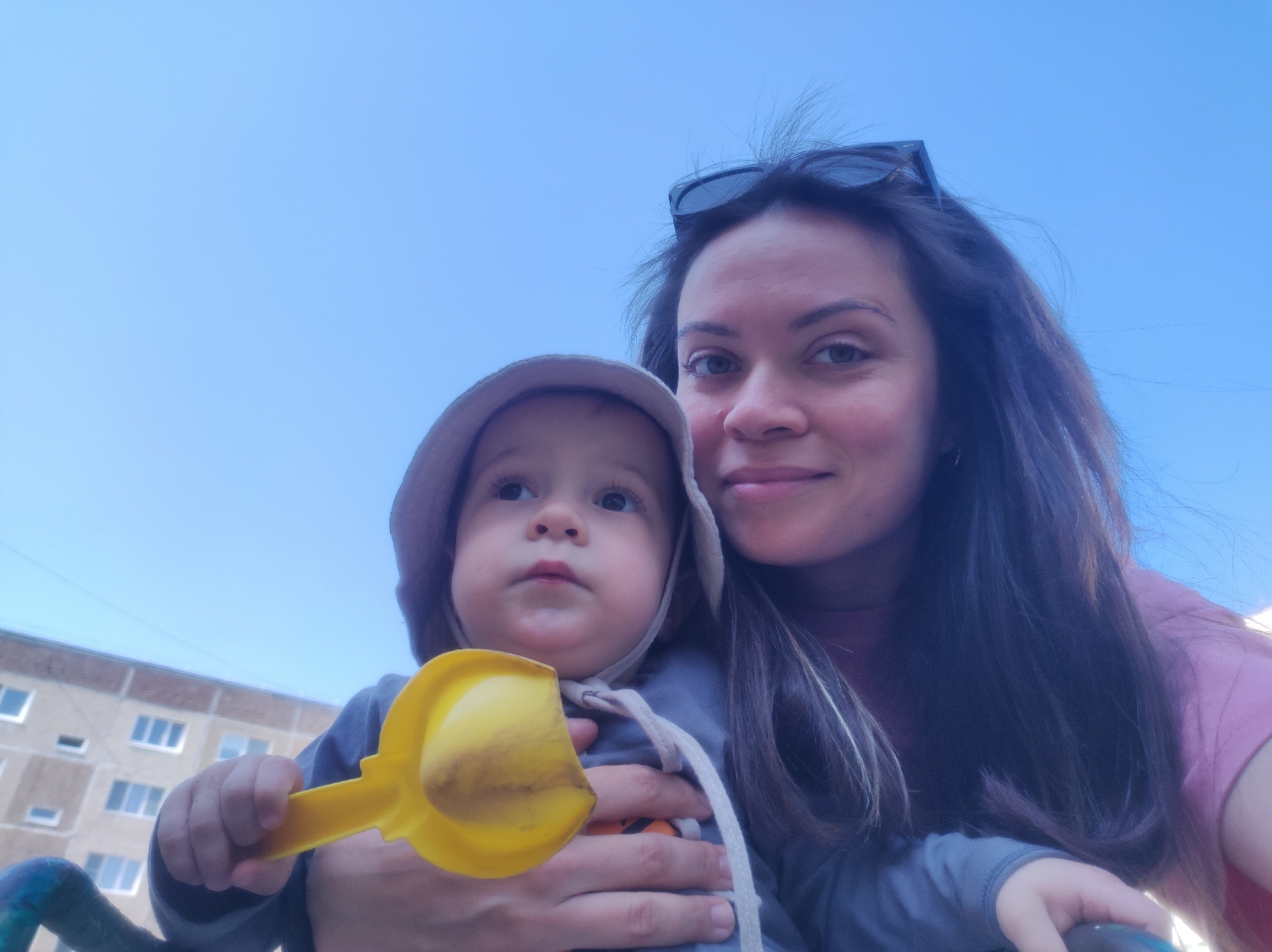 A selfie of a woman and a baby, likely taken in Chaikovsky, Russia, judging by the apartment block in the background. The woman has long dark hair, sunglasses on her head, and is wearing a pink t-shirt. She is smiling slightly. The baby is wearing a grey hat and a grey long-sleeved shirt and is being held by the woman. The baby is holding a yellow plastic toy scoop. The background shows a pale blue sky and a residential building with multiple windows. The bottom of the frame includes a blurred blue railing.