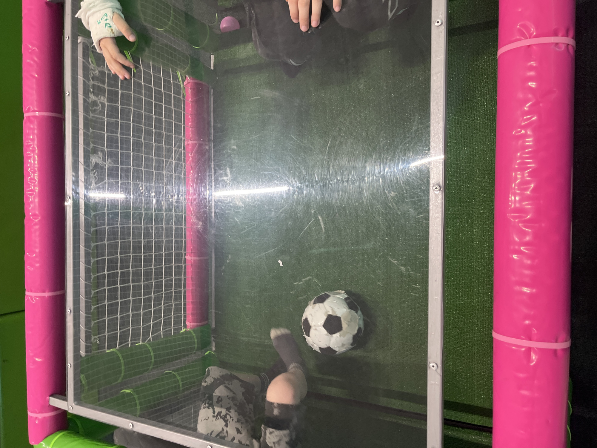The image depicts a foosball table inside a play area. The table features pink padded uprights and transparent walls. A black and white soccer ball is positioned near the bottom of the playing field. One player, wearing camouflage pants and a grey sock, is about to kick the ball. Another player is standing above the table, their hands visible. A third player with a bandaged hand is near the opposite goal. The goal is comprised of netting. The playing surface is green. The environment around the table consists of green padded flooring and walls. This activity appears to be taking place in a children's play center in Ulyanovsk, Russia.