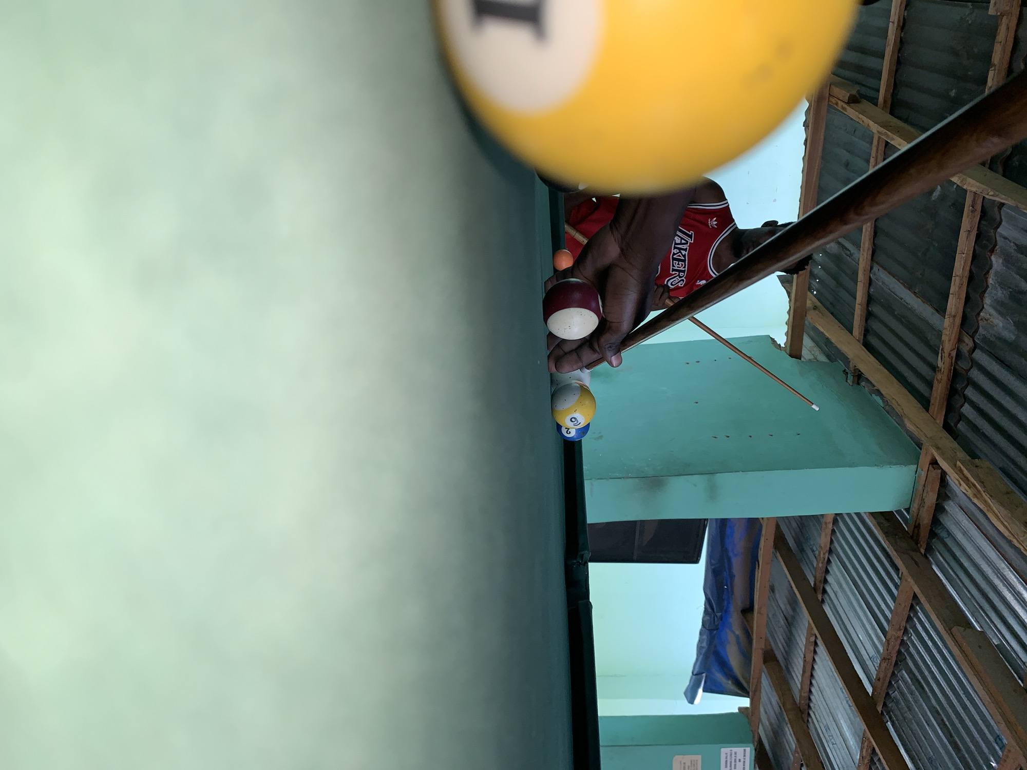 A man is seen playing pool on a billiards table. The table is covered with a light green felt and has three balls arranged on it. He is wearing a red Lakers jersey. The environment appears to be an informal pool hall with exposed beams and corrugated metal roofing. The location is New Stadium, Nigeria.