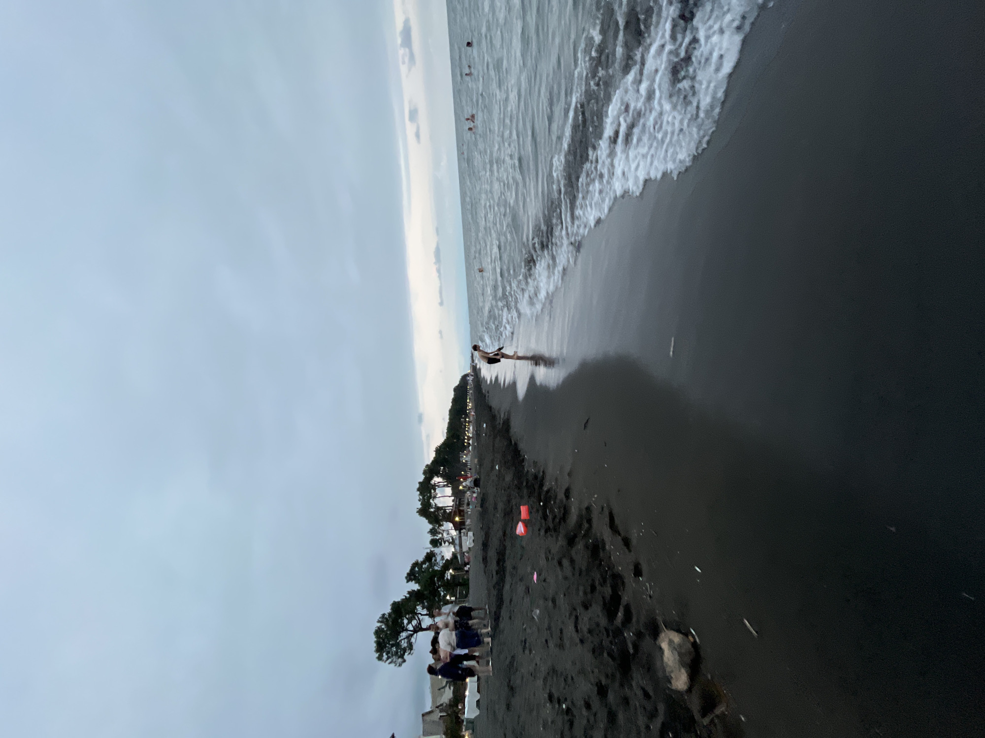 In Ureki, Georgia, a black sand beach scene unfolds under a cloudy sky. Several people are gathered on the shore, some standing in groups and others walking. One individual is near the water's edge, looking down. Further out in the sea, multiple people are swimming and enjoying the water. The shoreline is marked by the tide, with waves gently lapping at the dark sand. Scattered on the beach are some colorful plastic objects, possibly toys or debris. The background features a line of trees and structures along the coast.