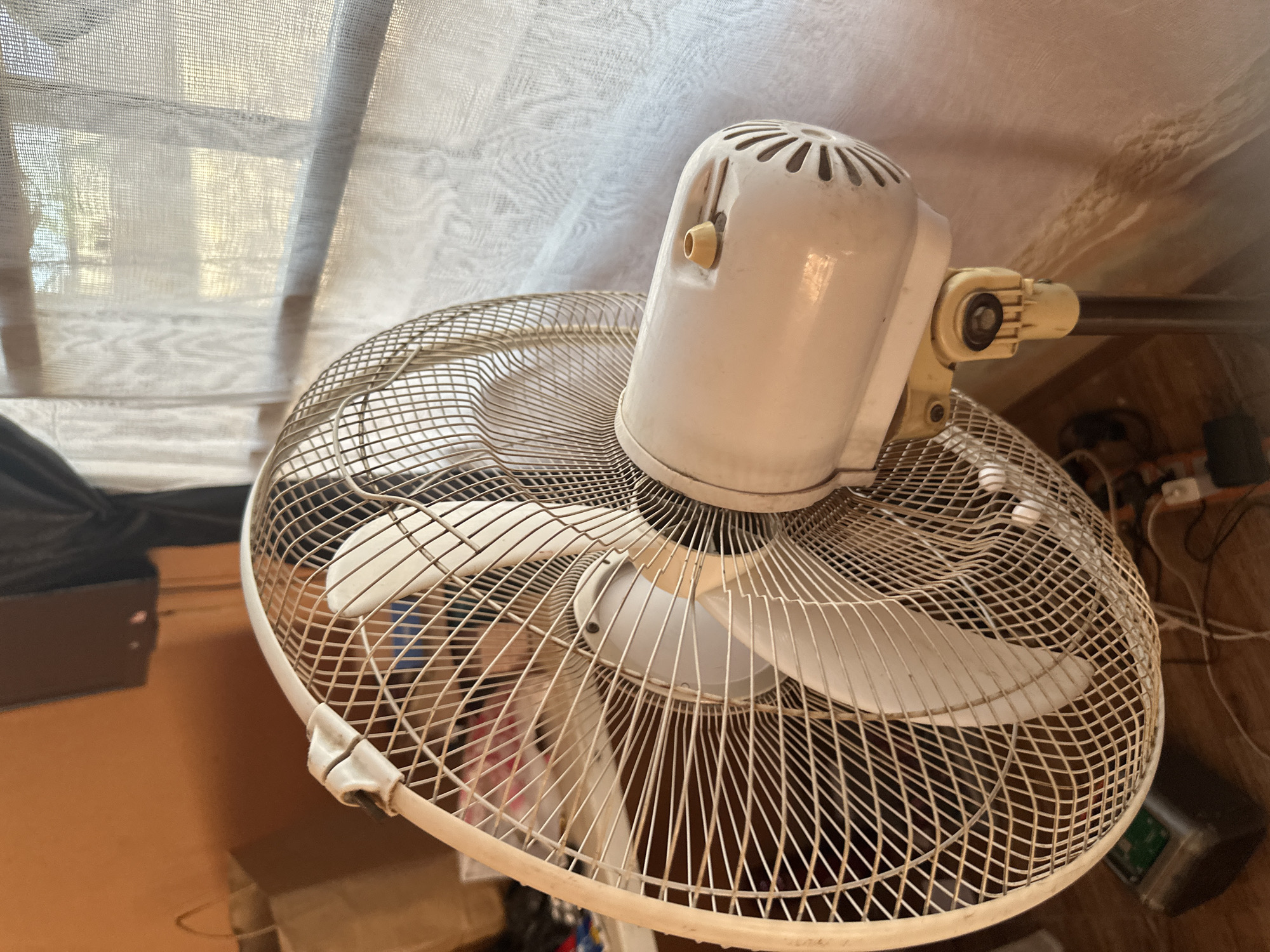The image shows a close-up, high-angle view of a white, vintage-style oscillating fan. The fan is in the foreground, dominating the frame. Its metal grill is a fine mesh, and the blades are white plastic. The motor housing is also white and has ventilation slits on top. A small, yellowish knob protrudes from the side of the motor housing, likely a speed control or on/off switch. The fan is positioned in front of a window covered by a sheer, white curtain, through which sunlight is filtering, creating a bright, diffused light. The background is blurred and suggests an indoor setting with wooden flooring and some electrical cords visible. There are no people or animals present. The overall impression is of a domestic setting, possibly a living room or bedroom, on a bright day. The fan appears to be stationary in this image, with no activity taking place other than its presence as an object. The context provided indicates the location is Jalingo, Nigeria, which might imply a warm climate where a fan would be in common use. No text is visible in the image.