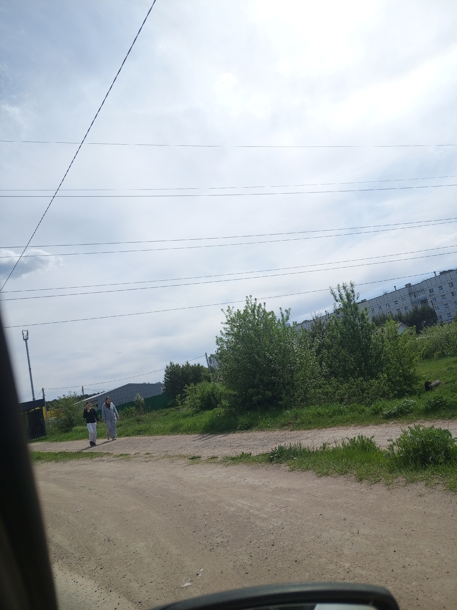 The photo, taken from a vehicle, captures a dirt road in what appears to be a rural or semi-rural area of Chaikovsky, Russia. Two women walk along the road. The woman on the left wears a dark top and light pants, while the other wears a light-colored tracksuit. The road is unpaved, and grass and weeds grow on either side. In the background, green shrubs and trees are visible, along with a multi-story building. Overhead are several power lines. A black section of the vehicle's interior is seen on the left and bottom of the frame. The sky is overcast with light clouds.