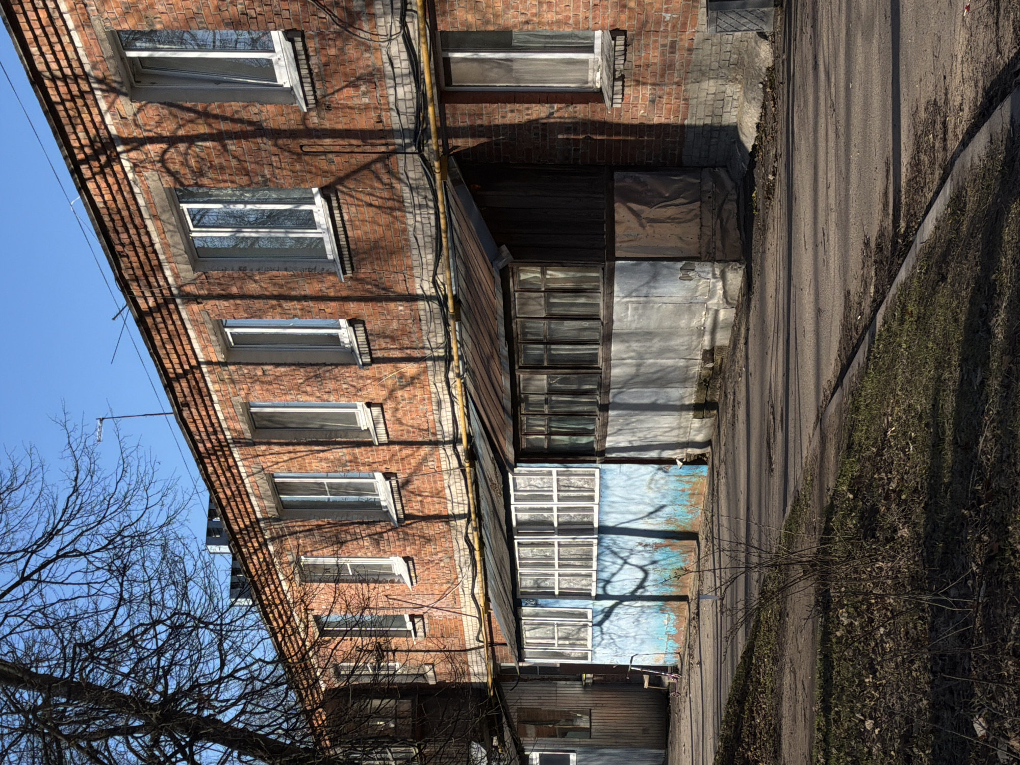 The image depicts a residential building in what appears to be Lavriki, Russia, based on the style and environment. The building has a brick facade, with several windows visible. A covered veranda or extension protrudes from the side of the building, with a mix of materials including wood, metal sheeting, and faded blue paint. The building faces a narrow paved road, with patches of dirt and dried vegetation along the edge. Bare tree branches are visible in the foreground, suggesting it might be late autumn or winter. No people are visible in the frame. The scene is bathed in bright sunlight, casting shadows of the trees and building on the facade. Electrical or utility lines run along the side of the building.