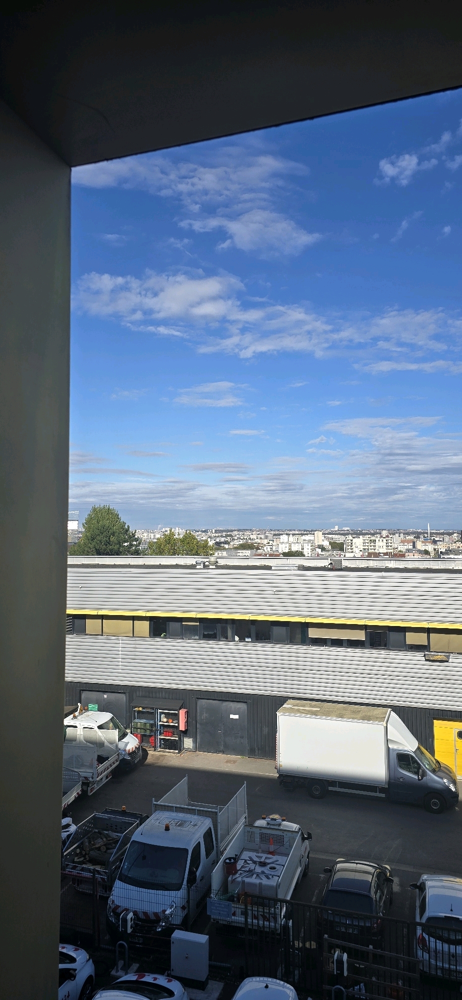 The image captures a view of an industrial area and a city skyline under a bright blue sky with scattered clouds. In the foreground, a building with a corrugated metal roof and yellow trim is visible, with several commercial vehicles parked in front of it. These vehicles include a white van with a trailer, a utility truck with a water tank, and another white van. Further in the background, a city skyline stretches across the horizon, dotted with buildings and trees. The scene is devoid of people. The overall impression is one of a functional, urban environment during daylight. The location is specified as Villejuif, France.