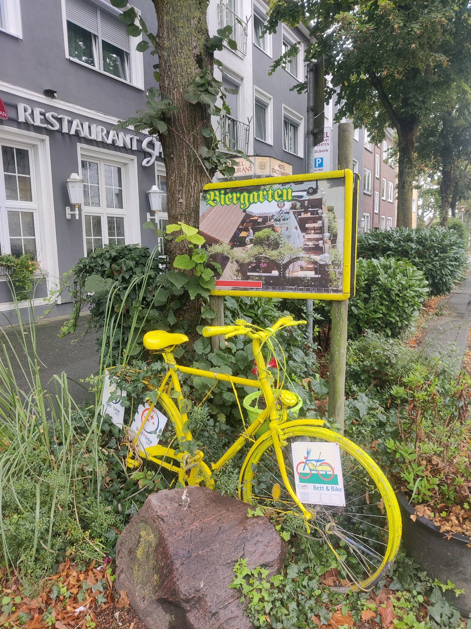 The image shows a bright yellow bicycle leaning against a tree on a sidewalk in Emmerich on the Rhine, Germany. The bicycle has a green pot attached to its handlebars and is surrounded by lush green foliage and fallen leaves. A large, rough-hewn rock rests in front of the bicycle.
Behind the bicycle, a yellow sign with the words "Biergarten" is prominently displayed. The sign features an illustration of people dining outdoors at tables under a canopy. The building in the background is a multi-story structure with grey siding and white-framed windows. A sign above the entrance reads "RESTAURANT". The sidewalk runs alongside a street lined with trees and shrubs.
A smaller sign with a bicycle and fork graphic is attached to the spokes of the bicycle's front wheel. The words "Bett & Bike" are printed below the graphic. A small black cat can be seen perched on the ledge of the restaurant building.