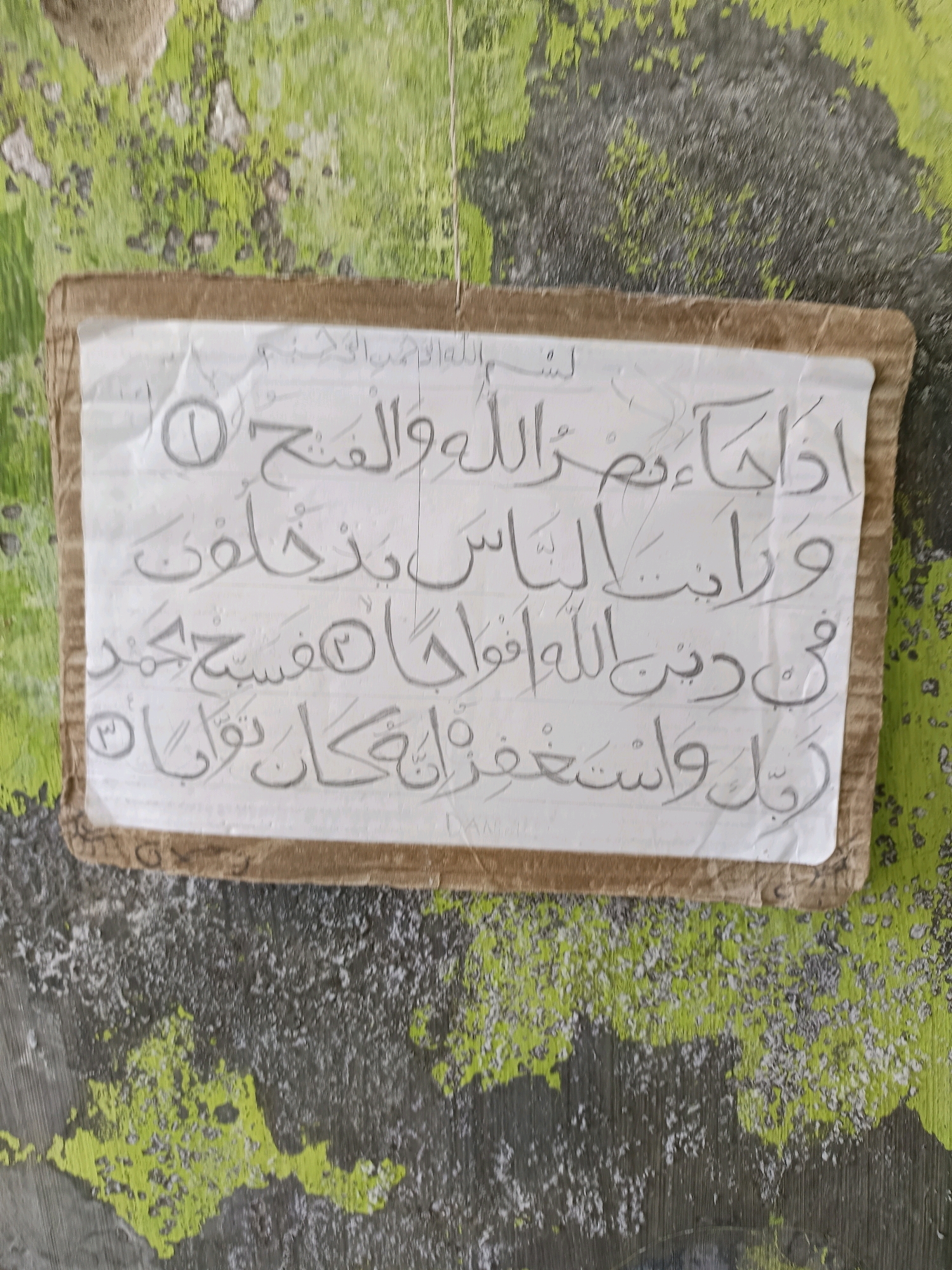 The image displays a handwritten Arabic text on a white paper, framed by brown cardboard. The text appears to be a religious verse or excerpt. The paper is affixed to a weathered wall with peeling green and grey paint, typical of an outdoor or semi-outdoor environment in Labuhan, Indonesia. The wall's texture is rough and uneven, showing signs of age and exposure to the elements. There are no people or animals visible in the image. The focus is solely on the text and its immediate surroundings.