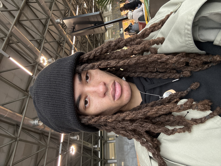 A male individual is depicted in a close-up, self-portrait style within Milan Malpensa Airport (MXP) in Somma Lombardo, Italy. The subject, with medium-dark skin, wears a black knitted beanie and has long, dark brown dreadlocks cascading over his shoulders and chest. He possesses dark eyes, defined eyebrows, and prominent lips, with visible stubble on his chin and upper lip. He is clad in a black hooded sweatshirt beneath a light beige or khaki trench coat. A silver-toned ball chain necklace with two circular pendants, one silver and one lighter in color, adorns his neck. The background reveals an indoor public area with a high ceiling composed of exposed metal beams, ducts, and multiple round, recessed light fixtures. To the right, a "Venchi" retail sign is partially visible, behind which several individuals are seated at tables within a lounge or cafe area. A large, dark, rectangular display screen stands in the mid-ground. The environment is brightly lit by artificial light sources.