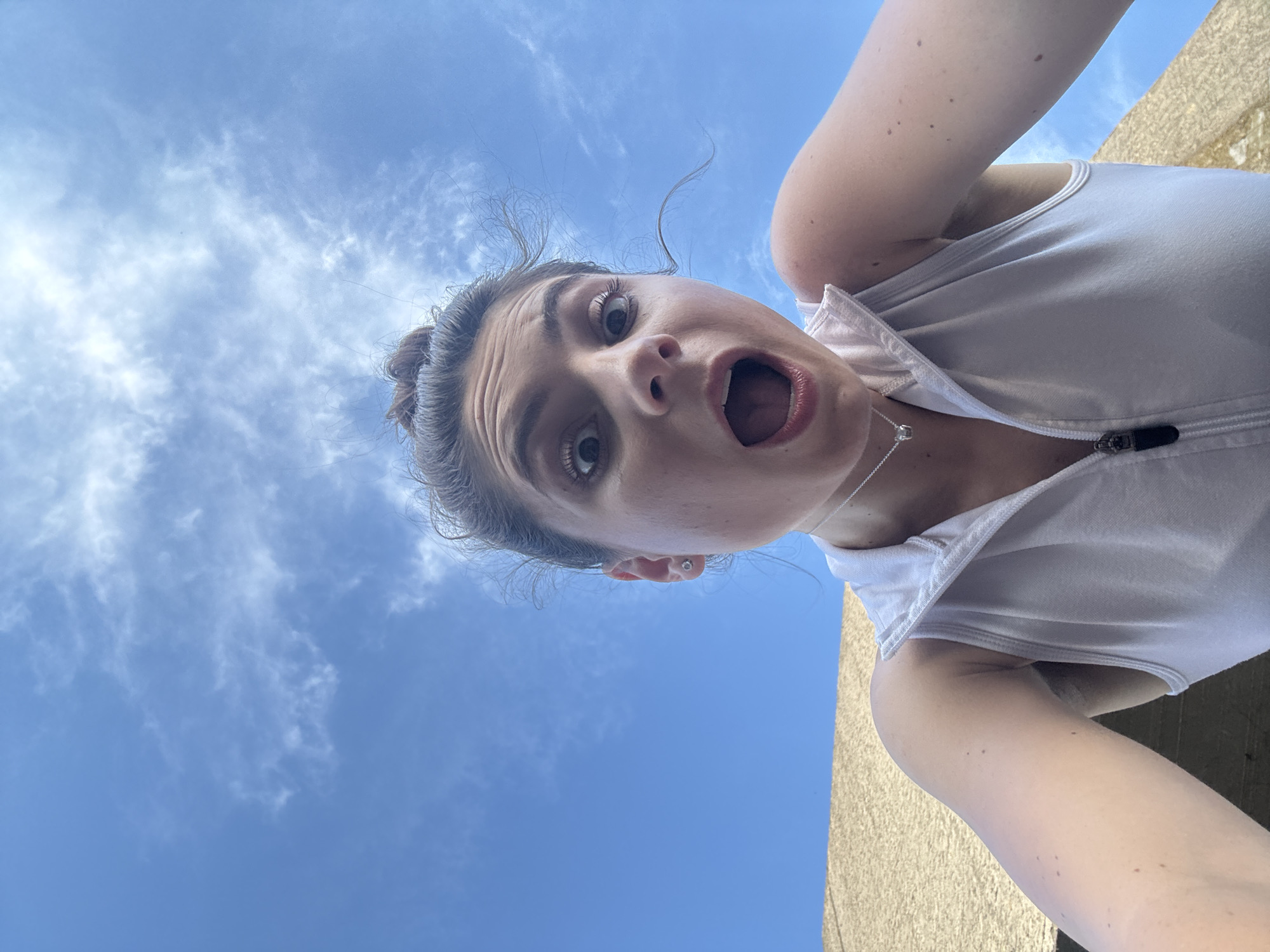 A woman with her hair in a bun and a surprised expression is looking into the camera against a blue sky with scattered clouds. She is wearing a white vest and a silver necklace. The lower part of the image shows a textured wall. The photograph was taken from a low angle, looking upwards. The woman's face is the primary focus of the image.