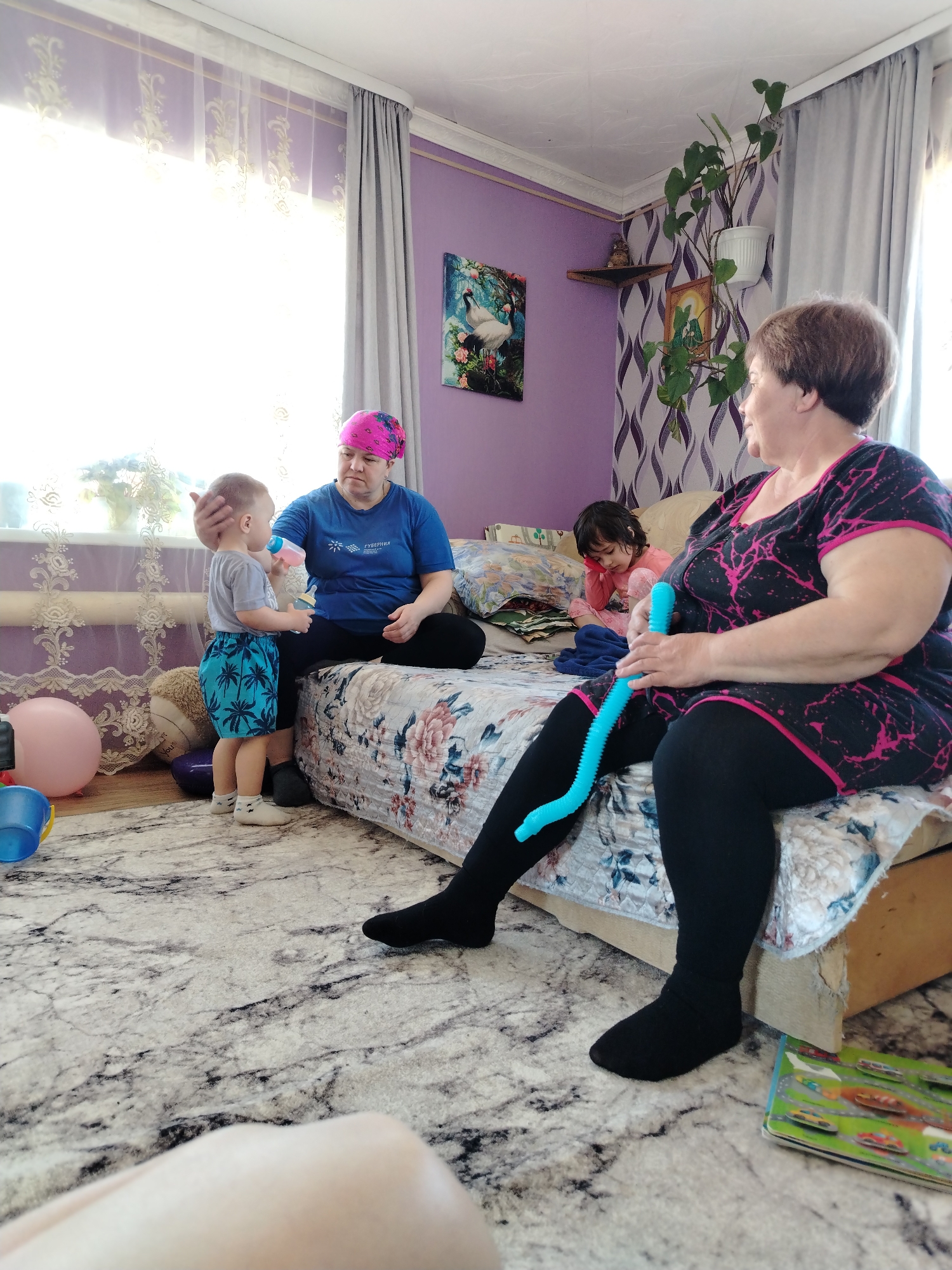 The image shows an interior scene. A woman sits on a bed with a floral bedspread, wearing a black dress with pink accents and black leggings. She holds a blue flexible tube toy. To her right, another woman wearing a blue t-shirt and a pink headscarf sits next to a young boy who is drinking from a bottle. A young girl is seated further back on the bed. There is a painting on the lilac wall behind them. A patterned rug is on the floor. A potted plant sits atop the window.