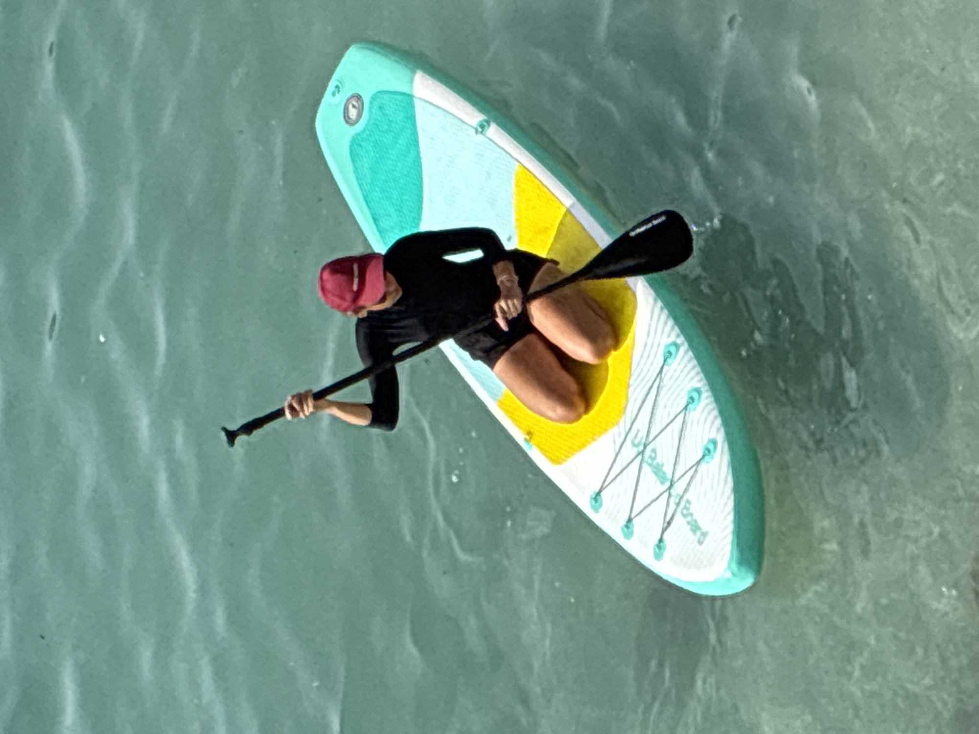 A person in a red cap and black long-sleeved rash guard and black shorts is kneeling on a paddleboard in the ocean. The paddleboard has a white, yellow, and teal design. The person is holding a black paddle and appears to be paddling. The water is a clear turquoise color with some ripples and small waves. The scene is likely a recreational activity in the waters off the coast of Alanya, Turkey, given the clear blue waters commonly found in the Mediterranean Sea in that region.