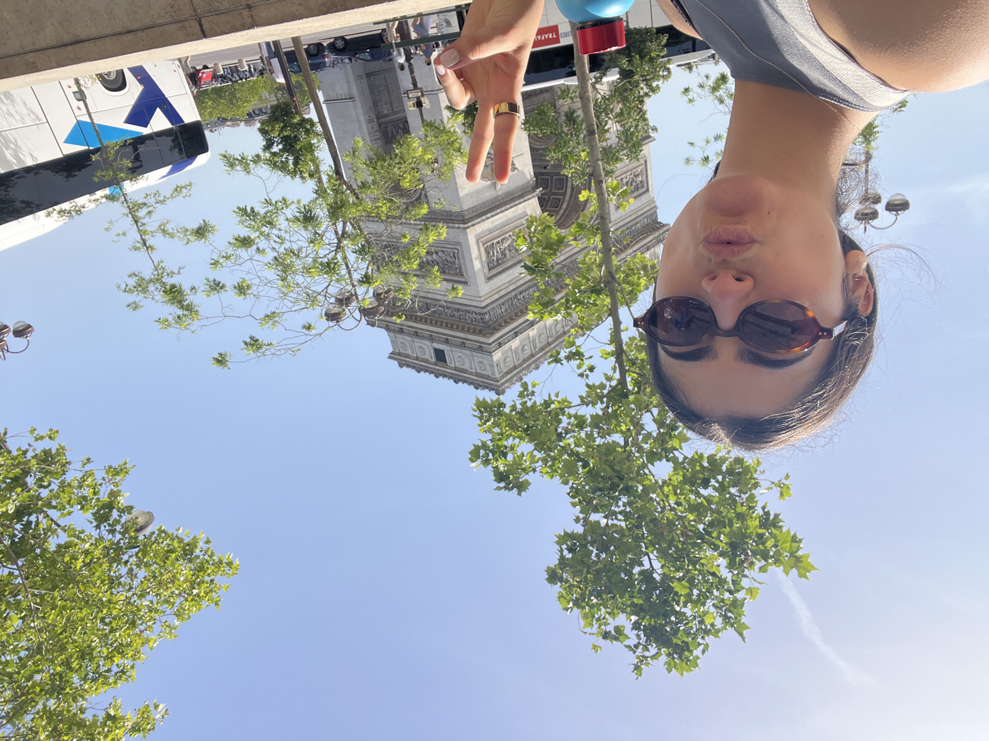 The photo shows a person in sunglasses posing with a peace sign in front of the Arc de Triomphe in Paris, France. A bus and other vehicles are visible in the background. The sky is blue with trees framing the monument.