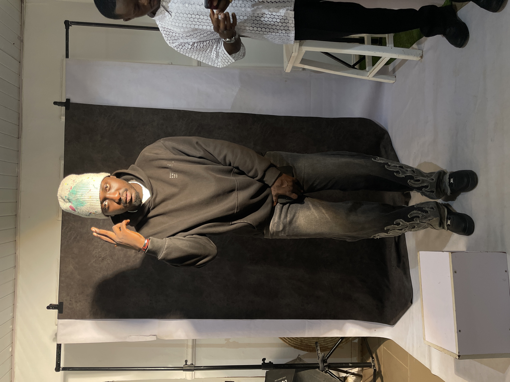 The photo shows a man in Barakin Akawo, Nigeria, standing in front of a photography backdrop with a dark grey background above a white section. The man is wearing a brown hoodie, patterned jeans, black shoes, and a beanie. He is looking at the camera and making a hand gesture. Above and behind him is another man wearing a white patterned shirt and black pants, standing on a small white stool and using a cell phone. In the background are lighting equipment and parts of a woven basket. To the right of the man in the hoodie is a small, white, square box. The scene is indoors.