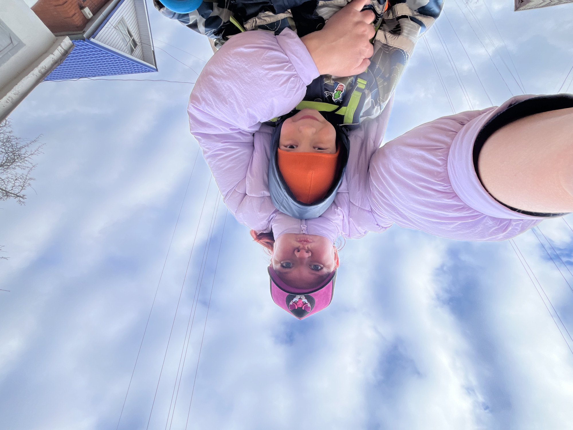 This unique photo captures two individuals, likely a child and an adult, from a worm's-eye view, giving the impression that they are hanging upside down against a cloudy sky. The camera appears to be placed on the ground or held low, with the subjects looking directly down into it.
The primary subject on the left is a young child wearing an orange knitted beanie under a grey or black hooded jacket, topped with a light lavender puffer jacket. A camouflage-patterned item, possibly a backpack or harness with green straps, is visible behind them, featuring a small patch depicting a cartoon radish. The child looks towards the camera with a neutral expression.
To the right, partially visible, is another individual, likely an adult, also wearing a light lavender puffer jacket. They have a pink baseball cap with a dark, intricate emblem on the front, and reddish-brown hair peeks out from under the cap. Their expression is also neutral, with a slight pout.
The background shows a pale, overcast sky with scattered white clouds. Several thin wires, likely power or communication lines, crisscross the upper parts of the frame. In what would be the top-left corner if the image were re-oriented normally, parts of residential buildings are visible: one with a dark orange tiled roof and white siding, and another with a distinctive blue shingled roof and a white facade featuring a window. Bare tree branches are also visible near these structures.
The image conveys a playful moment, suggesting the individuals are either hanging upside down from a structure like playground equipment or posing in an inverted position, creating a dynamic and unconventional perspective. The scene takes place outdoors during the daytime under cloudy weather, in a residential area, likely in Chaikovsky, Russia, as per the location context.
