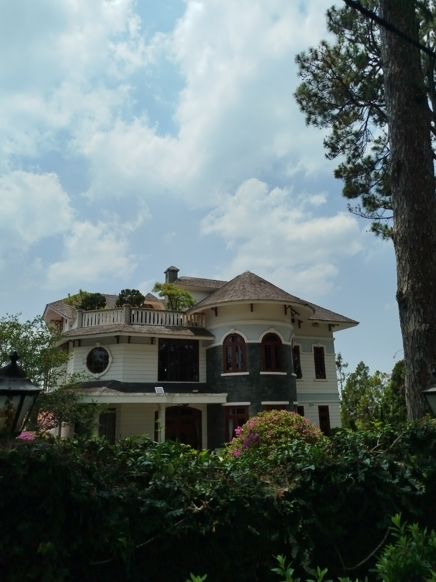 A daytime, eye-level shot features a large, ornate house partially obscured by dense, overgrown foliage, possibly a hedge or garden. The house has a multi-tiered structure with varying rooflines, including a rounded turret-like section. Architectural details include arched windows with dark frames, white clapboard siding, and a dark green accent wall. A rooftop terrace is visible, adorned with vegetation. A tall tree trunk dominates the right side of the frame, partially obscuring the house. The sky is a bright blue with scattered white clouds. The overall setting suggests a well-maintained property in a lush, possibly urban environment. This building is located in the city of Da Lat, Vietnam.