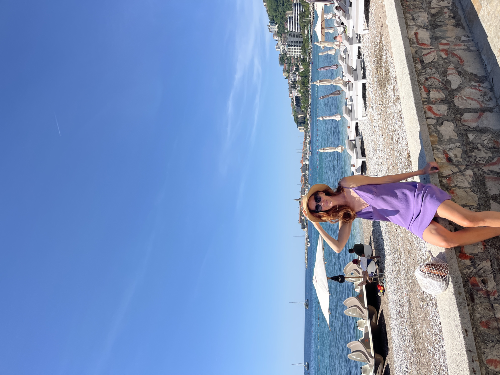 A medium-shot photograph shows a woman wearing a purple dress, sunglasses, and a straw hat resting on a concrete barrier next to a beach in Budva, Montenegro. The woman is positioned in the right portion of the frame, her hand resting on her hat and her body leaning on the barrier. To her left, there are beach chairs and a white umbrella positioned on a pebble beach next to the Adriatic Sea. Beyond the beach, the cityscape of Budva is visible, with buildings lining the coast and trees on the hills. A bright blue sky fills the left portion of the frame.
