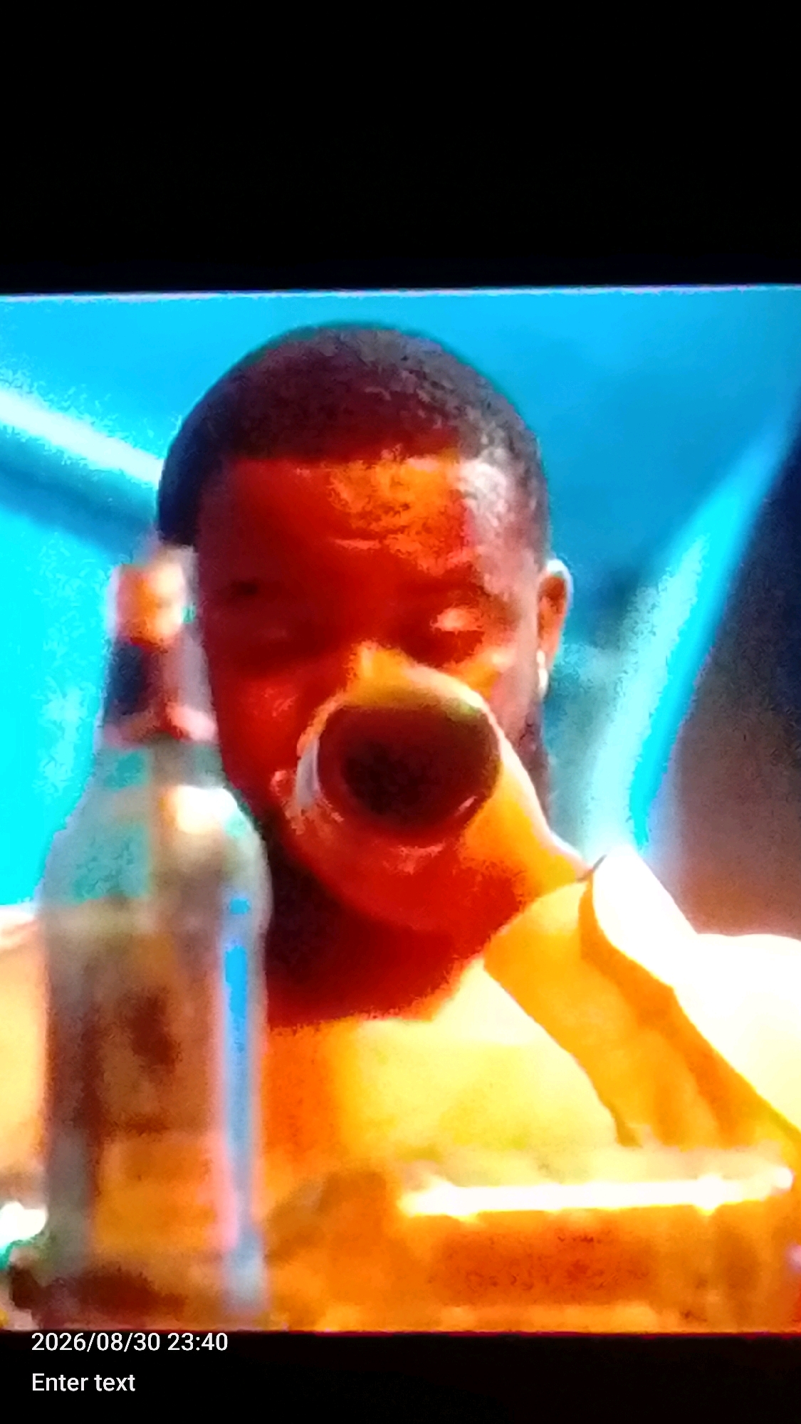The image captures a close-up, slightly blurry view of a man drinking from a bottle. The man's face is illuminated with warm, orange and yellow light, suggesting artificial lighting or a sunset. He has dark skin and short hair. In his right hand, he is holding a bottle, possibly a beer bottle, and he appears to be taking a sip. His left hand is near his mouth, holding a light-colored object, possibly the neck of the bottle or another item.
In the background, there is a vibrant blue color, which could represent a wall, a sky, or even water. The overall scene is abstract due to the lighting and focus, making it difficult to discern specific environmental details. The bottom of the image displays a timestamp "2026/08/30 23:40" and the text "Enter text", indicating that this is likely a screenshot or a captured image from a video or digital display.
Due to the limited visual information, it is impossible to pinpoint the specific location within Lokoja, Nigeria. The scene could be indoors or outdoors, in a residential area, a bar, or a recreational space.