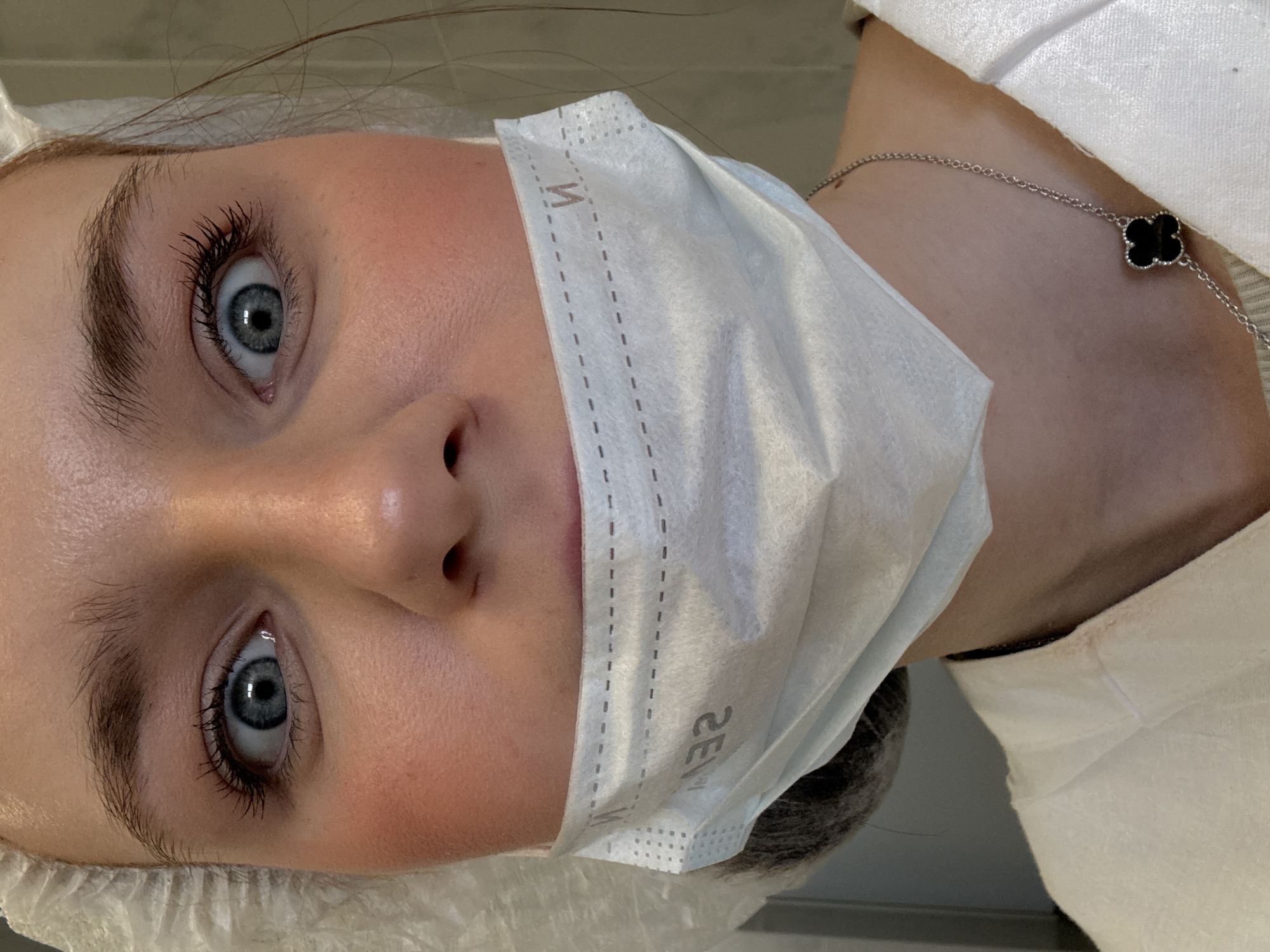 A close-up, slightly angled headshot of a person wearing a white surgical mask and a white disposable hairnet. The person has fair skin, light blue eyes with dark lashes, and natural eyebrows. A delicate silver necklace with a black four-leaf clover pendant is visible on their neck. The mask covers the nose and mouth, with the upper edge displaying a perforated design. The background is out of focus. The location is not discernible from the image.