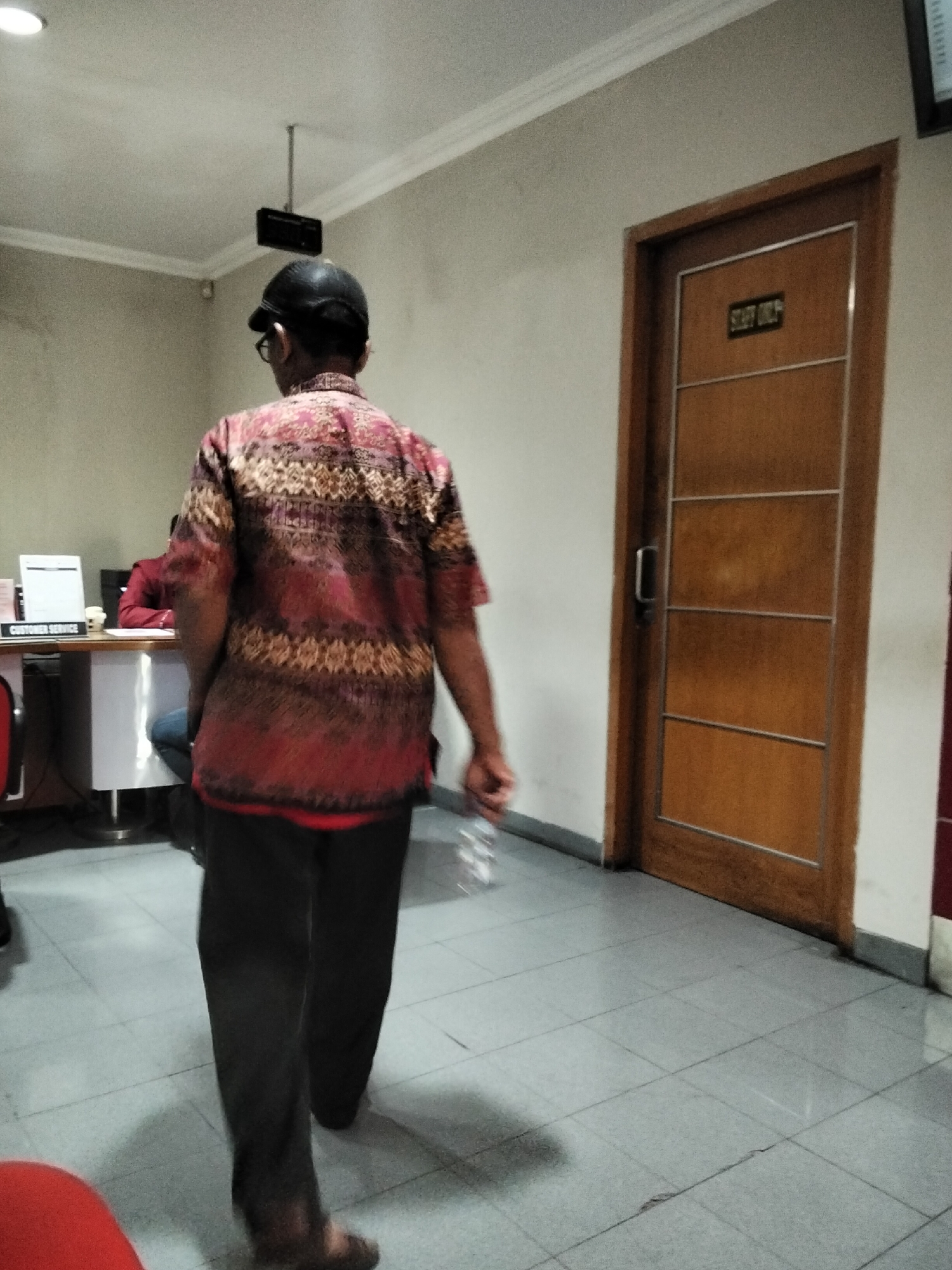 A person wearing a black baseball cap, glasses, a patterned short-sleeved shirt in shades of red and brown, and black pants is walking away from the camera in an indoor environment. The person is holding a clear plastic bottle in their right hand. To the right of the person is a wooden door with four horizontal panels and a metal handle. Above the door, a small sign reads "STAFF ONLY". In the background, to the left, is a desk labeled "CUSTOMER SERVICE" with a computer monitor and papers on it. Another person is partially visible behind the desk. The flooring is tiled in a grey checkerboard pattern. The walls are painted in a light, neutral color. The ceiling has a light fixture and a small black box mounted from the ceiling. This scene is likely located in a public service building or office in Kediri City, Indonesia.