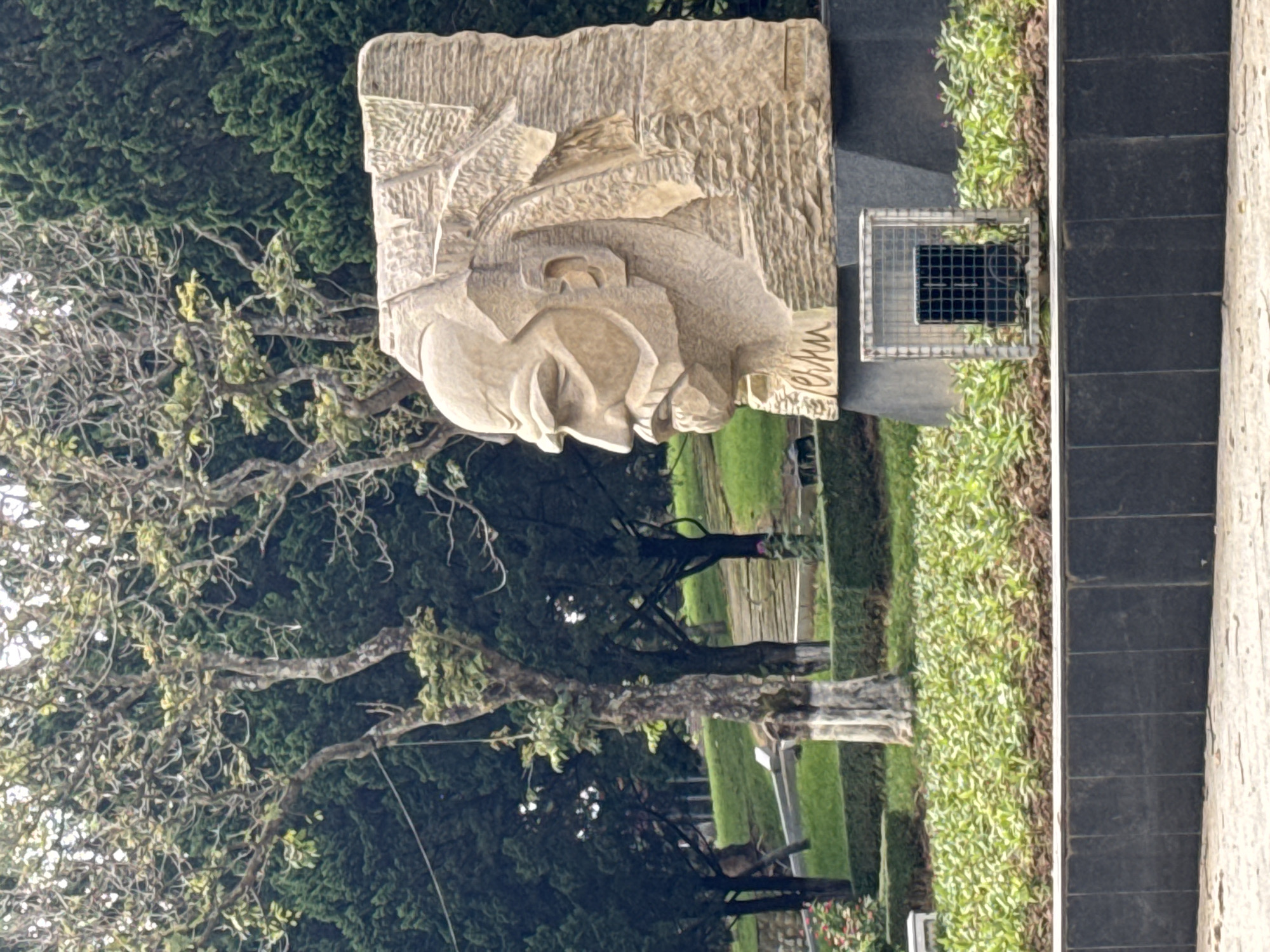 A large, stylized stone sculpture of Vladimir Lenin, depicted in a left-facing profile with a prominent beard and mustache, stands on a dark, multi-faceted pedestal in a park in Da Lat, Vietnam. The name "Lenin" is carved into the base of the sculpture. Below the sculpture, a small square metal cage encloses a dark rectangular object.
The immediate foreground features a dark, tiled retaining wall, beyond which lies a raised planter bed filled with numerous small, light-green leafy plants. The midground is characterized by manicured green grass and low, trimmed hedges. Further back, a paved path or set of steps ascends a gentle slope. The background is dominated by a dense arrangement of trees, including tall, slender trees with mostly bare or sparsely leafed branches, and darker, fuller coniferous trees. The lighting is soft and diffused, indicating an overcast sky or indirect light. No discernible people or specific actions are visible within the frame.
