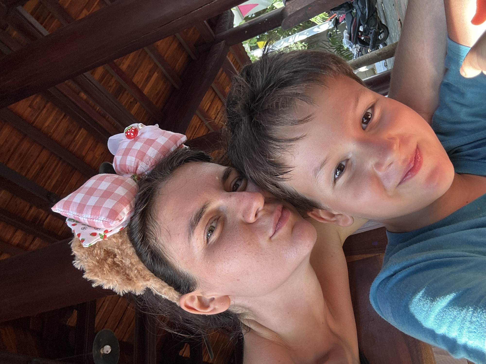 A selfie taken at an angle shows a fair-skinned woman wearing a headband with stuffed pink and white checkered cushions and a young boy in a blue t-shirt. The woman has long dark hair, freckles, and is looking at the camera with a neutral expression. The boy has short brown hair and brown eyes and is smiling slightly. Behind them is a roof of brown wood and what appears to be outdoor vegetation and possibly some equipment. The image was taken in the city of Mũi Né, Vietnam.
