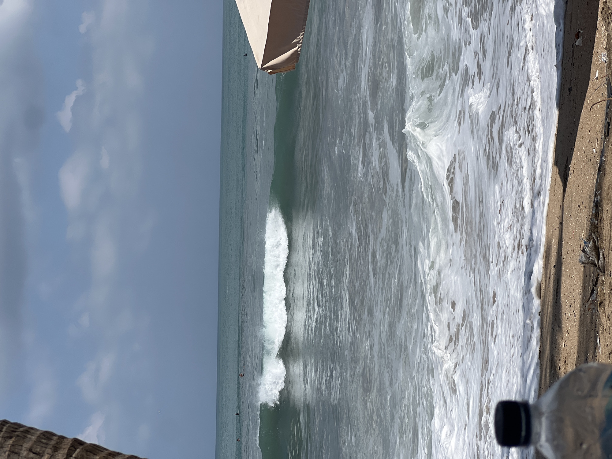 The image depicts a serene ocean scene with a gentle wave breaking in the mid-ground. The water is a tranquil turquoise, transitioning to a frothy white as the wave crests and collapses. Several surfers are visible as small figures in the distance, calmly waiting for the next wave. The sky above is a muted blue, adorned with scattered, fluffy clouds, hinting at fair weather. In the foreground, a sandy beach is partially visible, with a plastic water bottle in the lower left corner, suggesting a human presence. To the right, the corner of a tan beach umbrella provides a touch of shade. The overall atmosphere is one of peaceful coexistence between nature and recreational activity, capturing a typical day at the beach. This scene is highly characteristic of the popular Kuta Beach in Bali, Indonesia, known for its gentle waves and vibrant surf culture.