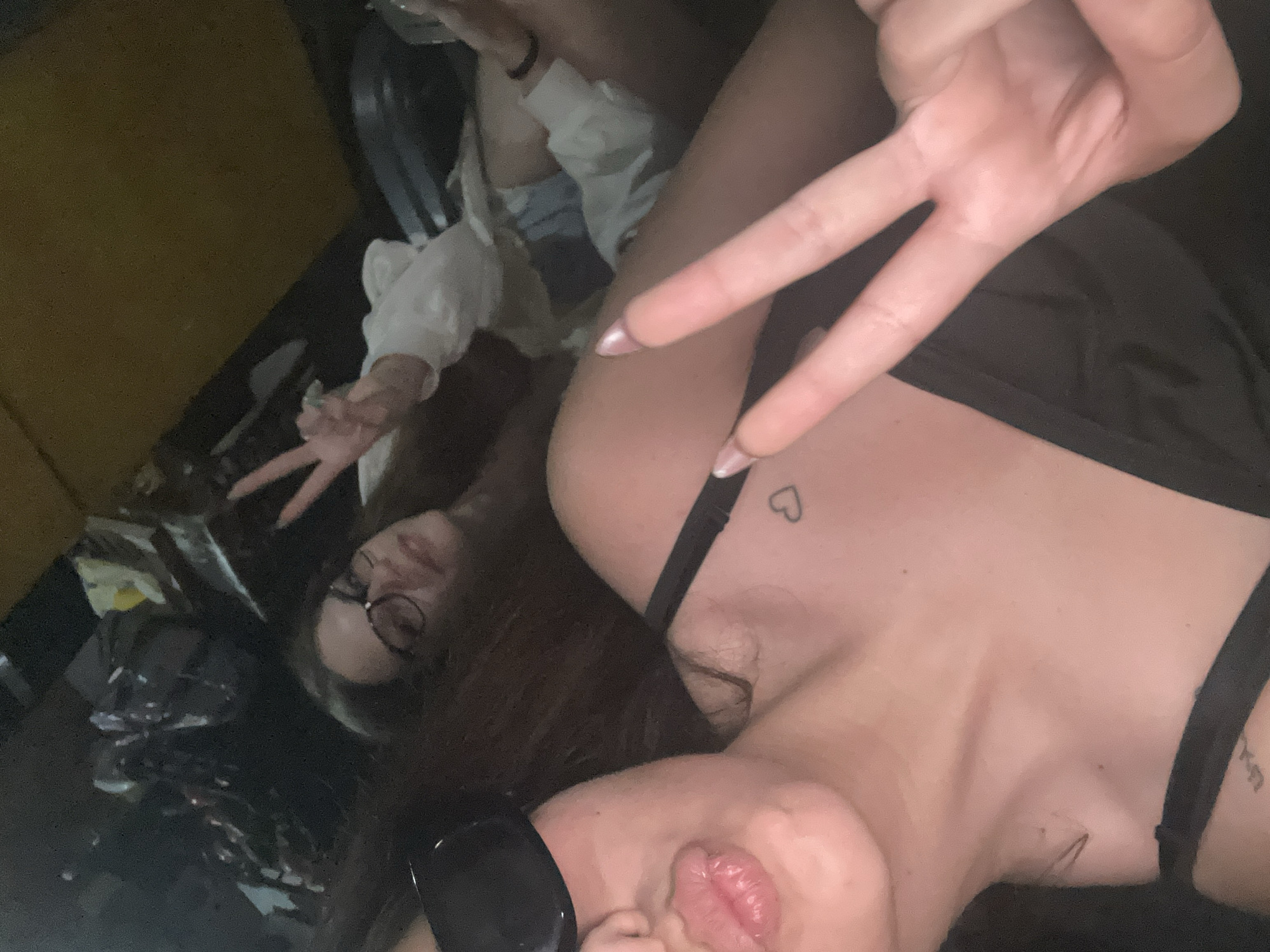 Two individuals, identified as young females, are depicted in a low-light, interior setting, consistent with the inside of a vehicle. The specific city and location are unknown.
The subject in the foreground, occupying the lower-left portion of the image, has long, dark hair. Her face is partially obscured by dark sunglasses covering her eyes. Her lips are prominent, pouted, and appear glossy with a light pink tint. She wears a black strapless top or bra. A small outline heart tattoo is visible on her left upper chest/shoulder area. Her left hand, extended towards the camera, displays a "peace" sign (V-sign) with two fingers, which have long, light-colored, possibly pearlescent, fingernails.
The second subject, positioned in the background and upper-right, has lighter brown hair and wears round-rimmed eyeglasses. She is smiling faintly and also displays a "peace" sign with her right hand. She is dressed in a white long-sleeved top with a slightly puffy sleeve design and light-colored denim shorts, with her legs visible.
The environment is dim, with artificial lighting casting soft shadows. The background features blurry dark shapes, possibly other seats or interior elements, and a section of yellow/gold upholstery in the upper-right corner. The overall perspective suggests a close-up selfie taken within an enclosed space.