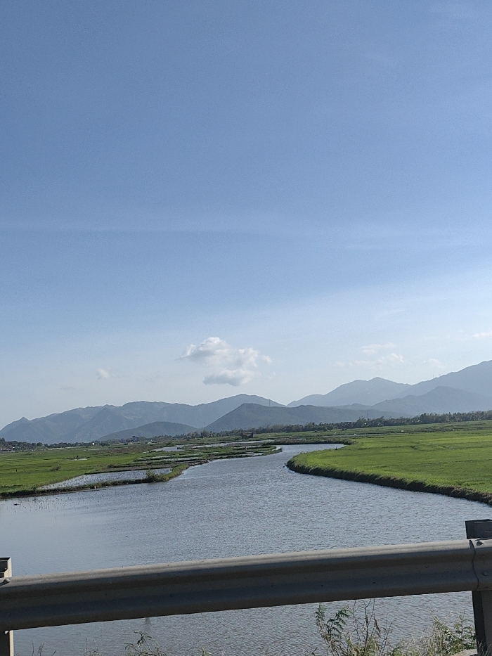 The image displays a landscape located in Ninh Hòa, Vietnam. A light grey metal guardrail, positioned in the immediate foreground, horizontally bisects the lower portion of the frame. Beyond the guardrail, a winding body of water, consistent with a river or large canal, flows from the center-left towards the right, then curves back towards the viewer. This water body is flanked by extensive, level agricultural fields, appearing vibrant green and partially submerged, indicative of rice paddies. In the mid-ground, the fields are segmented by visible embankments or smaller water channels. The distant background features a continuous range of blue-grey mountains, appearing hazy against the horizon. The sky is clear and bright blue, occupied by several scattered white cumulus clouds, one more prominent in the center-left. No individuals, specific actions, or overt structures other than the guardrail are discernible.