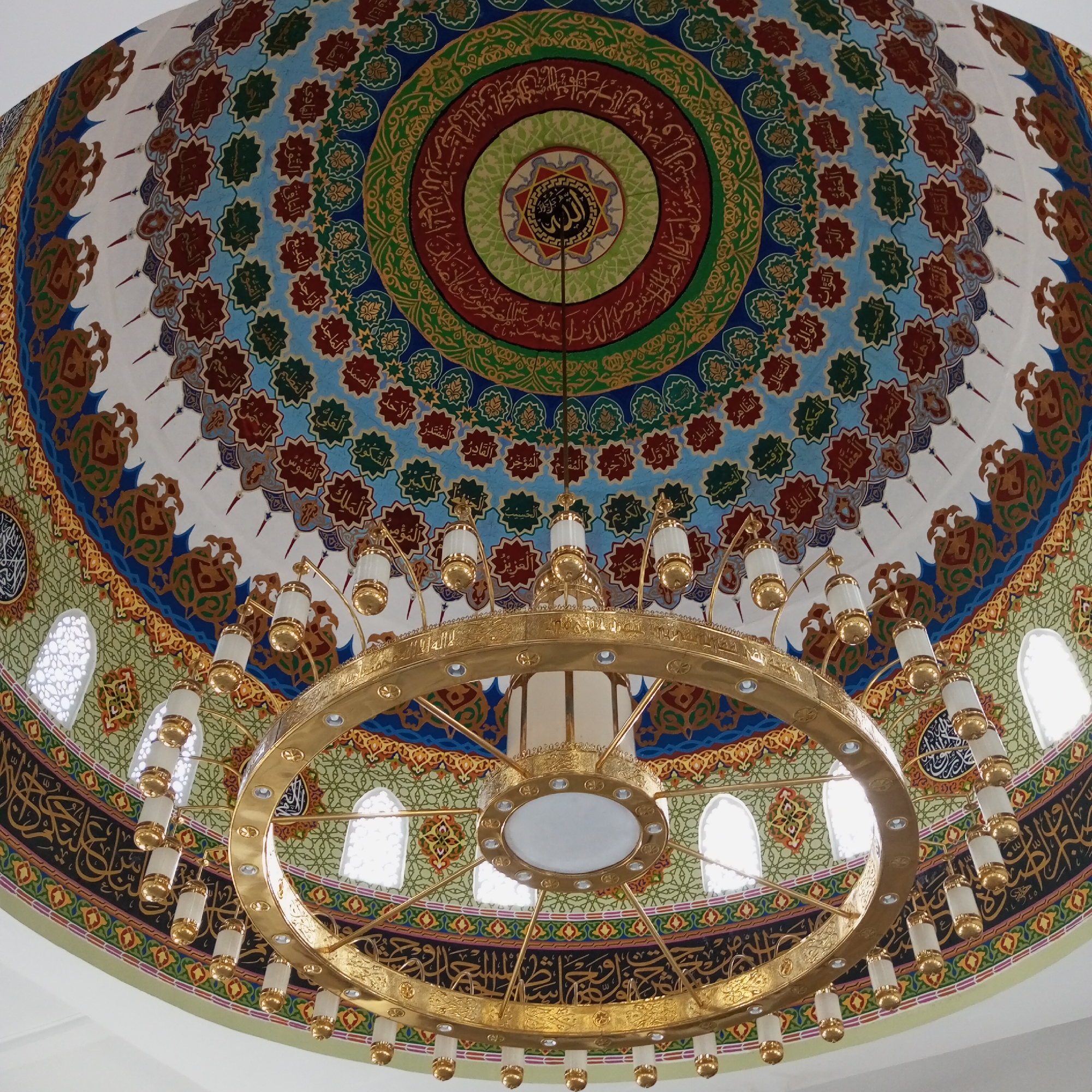 The image captures the interior of a mosque, specifically the dome, featuring ornate Islamic art and architecture. A large, golden chandelier is suspended from the center of the dome. The dome is decorated with concentric circles of intricate patterns and Arabic calligraphy. The color palette includes blues, greens, reds, and browns against a white backdrop. The chandelier has multiple tiers of lights. Windows are visible around the perimeter of the building, allowing natural light to enter the space. The location of this scene is identified as Meunasahaluedrienlhoksukon Barat, Indonesia.