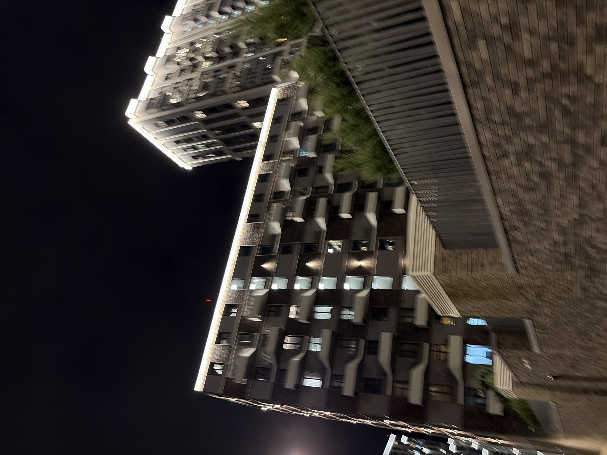 A night-time, low-angle shot captures the exterior of two modern apartment buildings in Moscow, Russia. The building in the foreground is a multi-story brick structure with balconies and numerous windows, many of which are illuminated. A bright white light runs along the top edge of this building. Behind it, a taller, sleeker building with a more complex facade, also illuminated by lights, rises into the dark sky. A dark metal fence with vertical slats runs horizontally across the middle of the image, separating the buildings from a brick wall in the foreground. Small, green trees are visible behind the fence, partially obscuring the lower portion of the taller building. The sky is dark and appears overcast, with no stars visible. The overall impression is one of urban development and modern architecture at night.