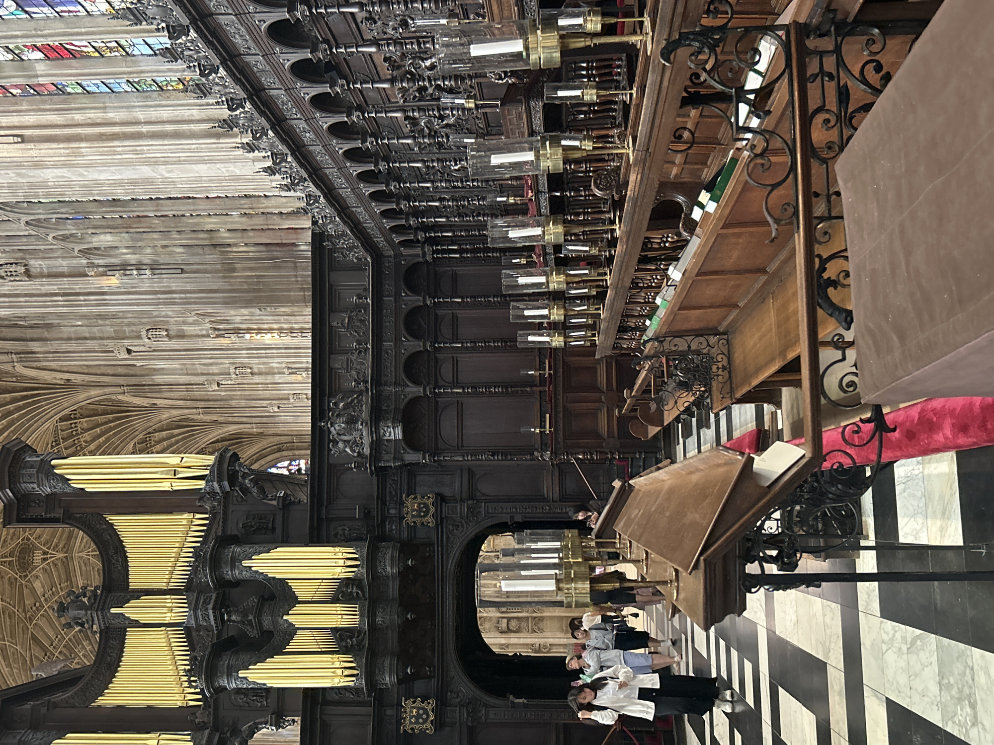 The image shows the interior of King's College Chapel in Cambridge, United Kingdom. The chapel features a grand organ with golden pipes on the left, intricate dark wood carvings throughout, and soaring vaulted ceilings. Rows of wooden choir stalls with ornate metalwork are visible on the right, illuminated by tall candelabra. In the foreground and mid-ground, people are seen walking on a black and white checkered floor. The scene appears to be during visiting hours, with several individuals dressed in casual attire observing the architecture. The atmosphere is one of awe and historical grandeur.