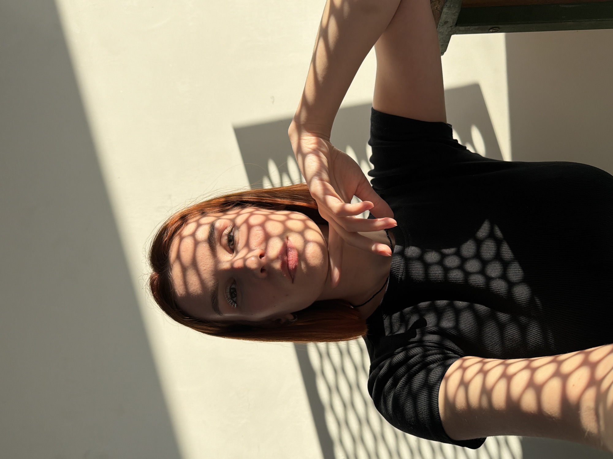 A portrait of a young adult female is depicted in an indoor setting in Chaikovsky, Russia. She is shown from the upper chest, directly facing the camera with a neutral expression. Her hair is reddish-brown, short, and straight. She wears a black ribbed short-sleeved top and a thin black choker around her neck. Her left hand is positioned under her chin, with fingers slightly curled, while her right forearm rests on a dark green, textured object, likely a chair component. Strong, directional sunlight casts prominent, uniform honeycomb-patterned shadows across her face, upper body, and the plain, light-colored wall behind her. A distinct diagonal shadow line is also visible on the upper section of the background wall.