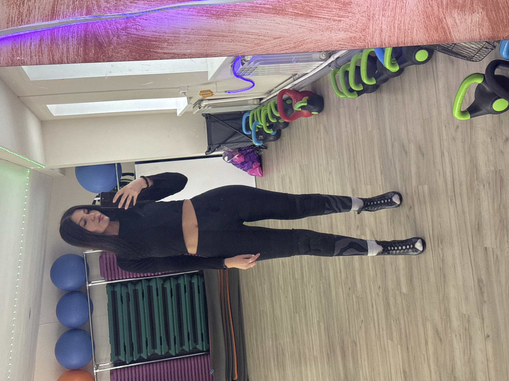 A woman in black athletic wear takes a mirror selfie in what appears to be a gym or fitness studio. She is standing in front of exercise equipment. The floor is a light-colored wood laminate. Weights and exercise balls are visible in the background. The location is uncertain, but the city is Chaikovsky, Russia.
