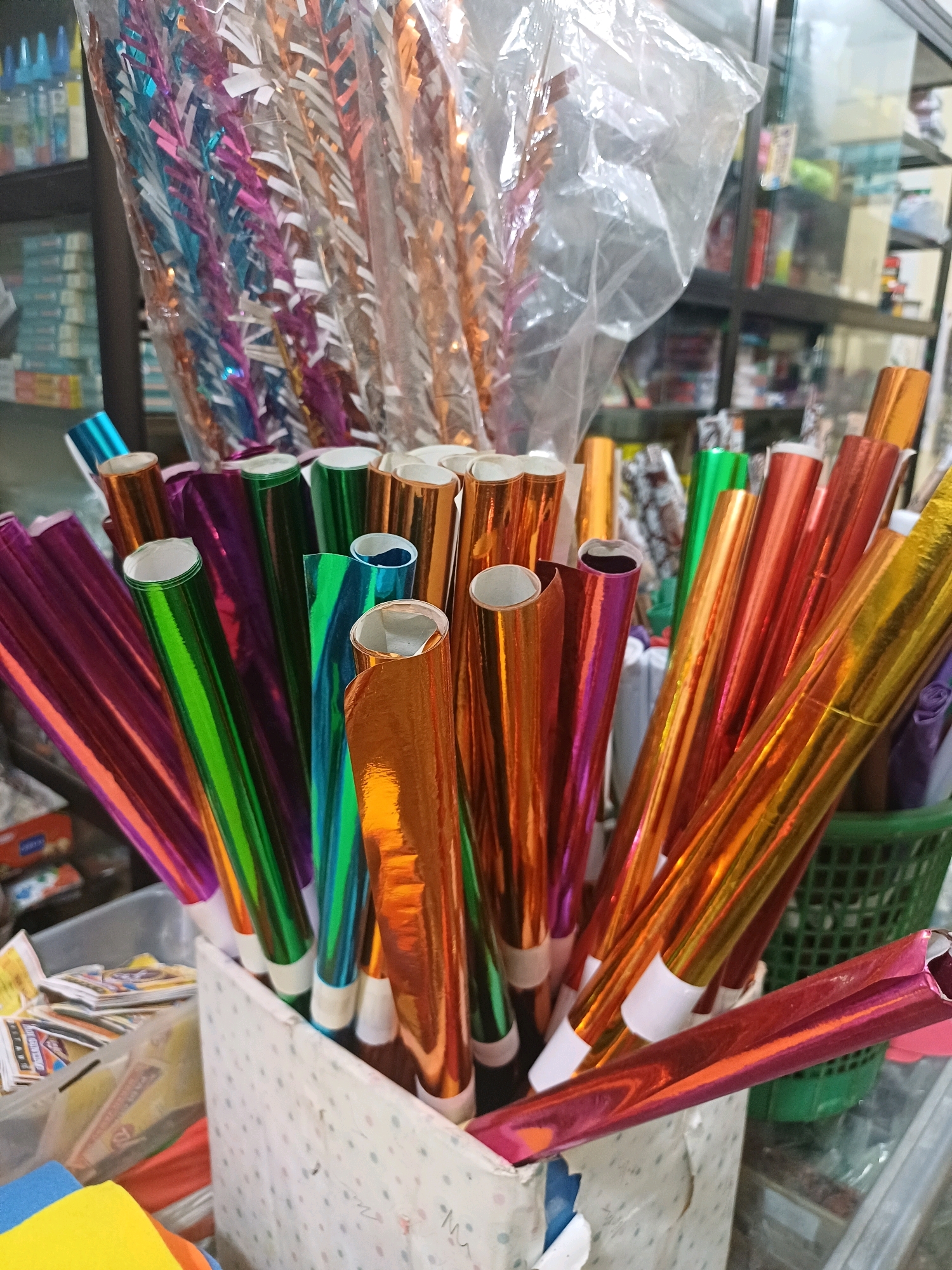 The image shows a close-up of various colorful metallic paper rolls, likely used for crafts or decorations. The rolls are primarily in shades of orange, green, pink, and purple, and they are bundled together in a speckled white box. Behind the box, a blurred background shows shelves stocked with various items, suggesting a retail or market setting. The overall impression is one of a colorful display of craft supplies. Without any visible landmarks or specific clues related to Dumai City, Indonesia, its precise location within the city cannot be determined. The scene is static, with no people or actions depicted.