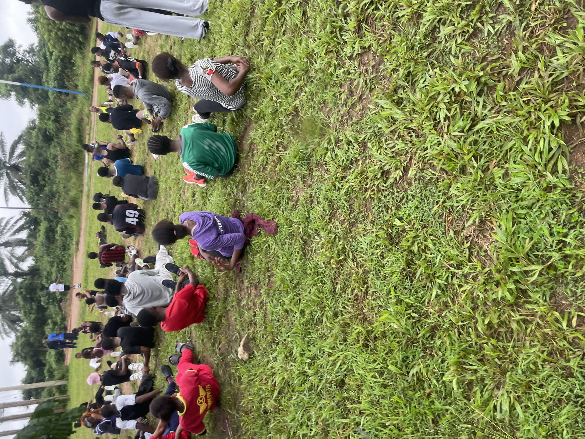 The image shows a group of young people sitting on a grassy field, likely engaged in an outdoor activity or training session. The setting appears to be a rural or semi-rural area, with lush green grass and trees in the background. The people are dressed in casual athletic wear, with some wearing brightly colored shirts.
The individuals are mostly seated, with some appearing to be in various stages of stretching or exercise. Their postures suggest they are listening to instructions or resting between activities. The presence of a blue and white pole in the background might indicate a sports field or training ground. The overall atmosphere seems to be one of communal activity and physical exertion.
The location is specified as Ihiagwa, Nigeria. This suggests the image was captured in a community within Nigeria, likely in an open area suitable for group gatherings and physical activities. The environment appears to be outdoors, with natural lighting and a backdrop of vegetation.