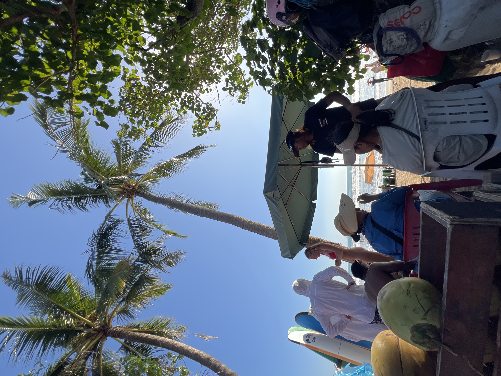 In Kuta, Indonesia, a beach scene unfolds under a bright blue sky, with two large palm trees framing the view. In the foreground, two green coconuts sit in a wooden cart. To the left, three surfboards are propped up, one with "BALI SURF" visible. A person wearing a white hooded jacket stands near the surfboards, facing away from the camera. Beside them, a woman in a red bikini converses with someone off-camera.
Underneath a green beach umbrella, a man wearing a black t-shirt and a baseball cap is standing. To the right, a woman with a straw hat sits in a white plastic chair, looking towards the ocean. Another person, wearing a light grey top and a pink helmet, is seated behind her. In the background, beach chairs and umbrellas are visible, along with a few people walking on the sand and in the water. The ocean is calm with small waves.