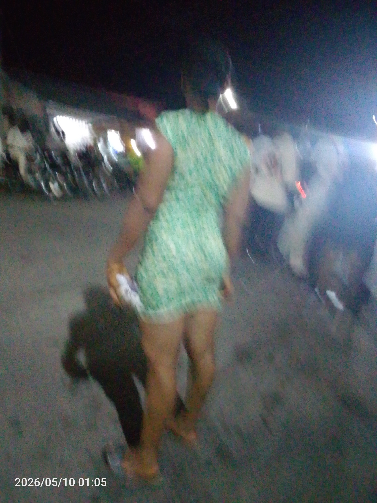 A medium shot, taken at night, depicts an individual in a short, green dress walking away from the camera. The setting appears to be outdoors, possibly a street in Lokoja, Nigeria, given the presence of parked motorcycles and other blurred figures in the background. The ground is paved, and the lighting is dim, with some bright spots causing flares in the image. The time stamp on the image reads 2026/05/10 01:05. The subject's shadow is visible on the ground.