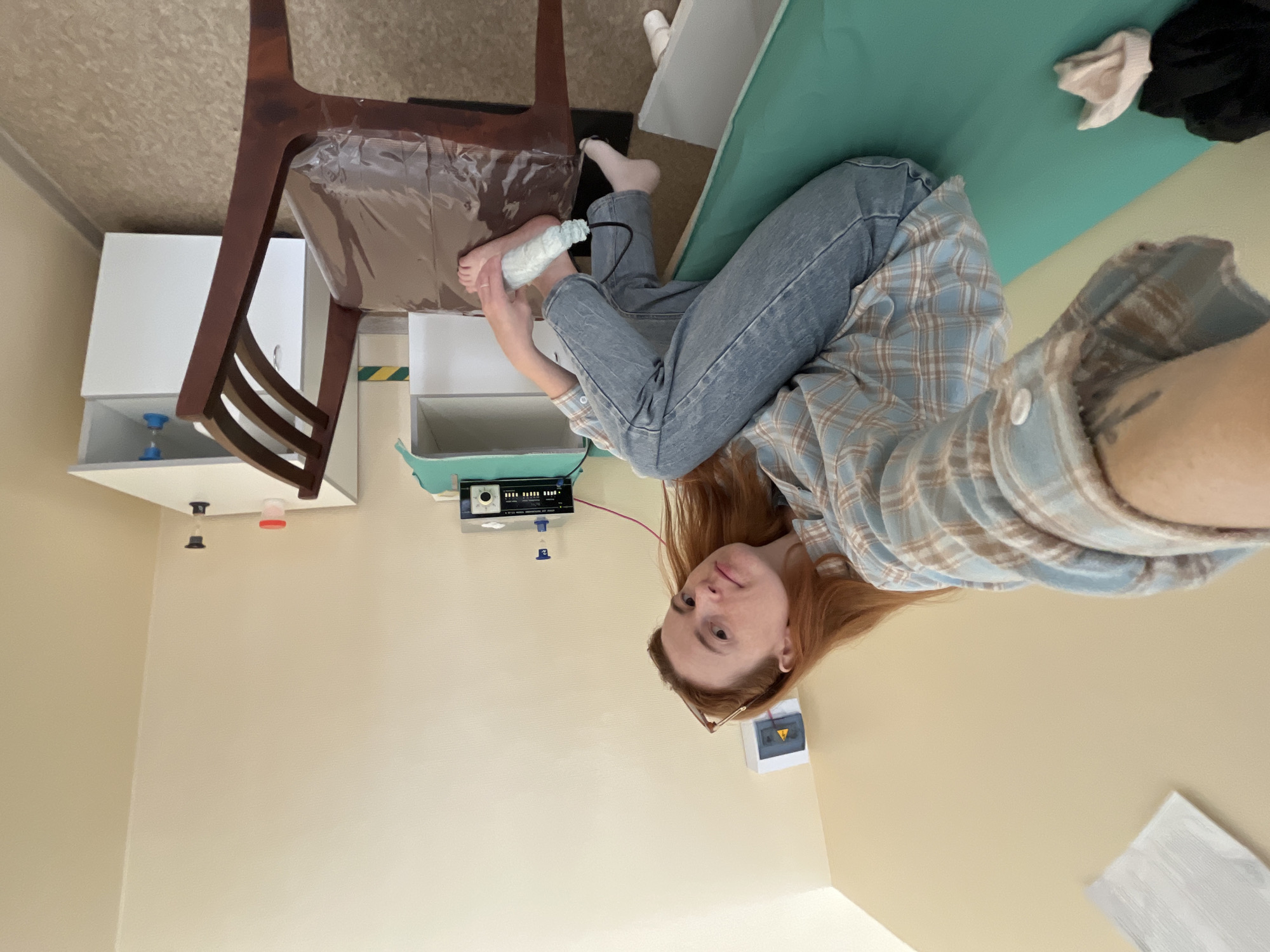The image, taken in Chaikovsky, Russia, features a young woman with long reddish-brown hair lying on a light green examination table or bed. She is wearing a light blue and brown plaid shirt and light blue jeans, looking directly at the camera with a neutral expression.
Her left foot is elevated, and a white, bandage-like wrapping is visible around her lower leg and foot, from which a black cable extends. This cable connects to a small, dark device resting on a white shelf, indicating a medical procedure, monitoring, or treatment related to her foot or ankle.
The setting appears to be a medical room or clinic. In the background, there is a dark wooden chair with a plastic-covered seat. Behind the chair are white cabinets with various items on the shelves, including a blue hourglass-shaped object and a small red-rimmed container. On a lower shelf, a black medical or laboratory device with several dials and indicators is visible, alongside a white plastic bottle with a blue cap and the number "2" printed on its side. The room has light-colored, possibly beige, walls. The lighting suggests an indoor daytime setting. No specific time of day or weather details are discernible from the interior view.