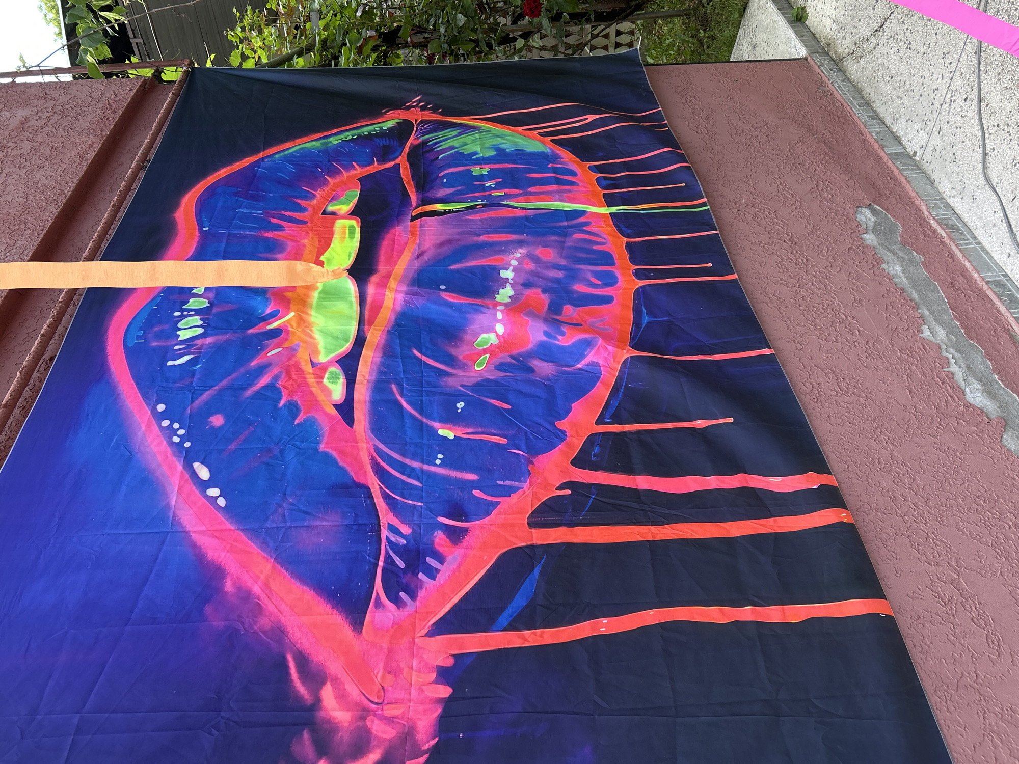 A close-up, eye-level, outdoor shot shows a large, dark blue tapestry featuring a vibrant, neon depiction of lips with dripping paint effects. The lips are outlined in bright pink and filled with lime green and electric blue. A vertical, bright orange streamer hangs from the top edge of the tapestry, partially obscuring the left side of the lips. The tapestry is affixed to a rough, reddish-pink textured wall. To the right of the tapestry, lush green foliage and the side of a red-tiled roof are visible, suggesting an outdoor setting. The ground beneath the wall appears to be a light-colored, textured patio.