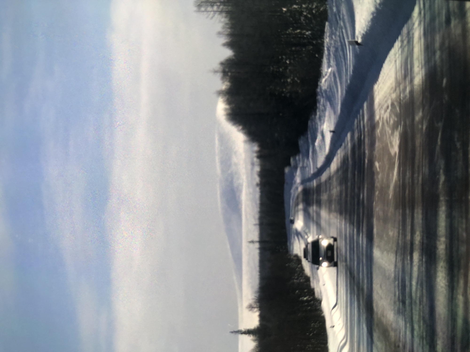 The image depicts a winding road covered in snow, bordered by a dark, dense forest on one side. A light-colored vehicle is traveling on the road. The sky is predominantly cloudy with patches of blue. The location appears to be a remote, possibly mountainous area. The city is unknown.