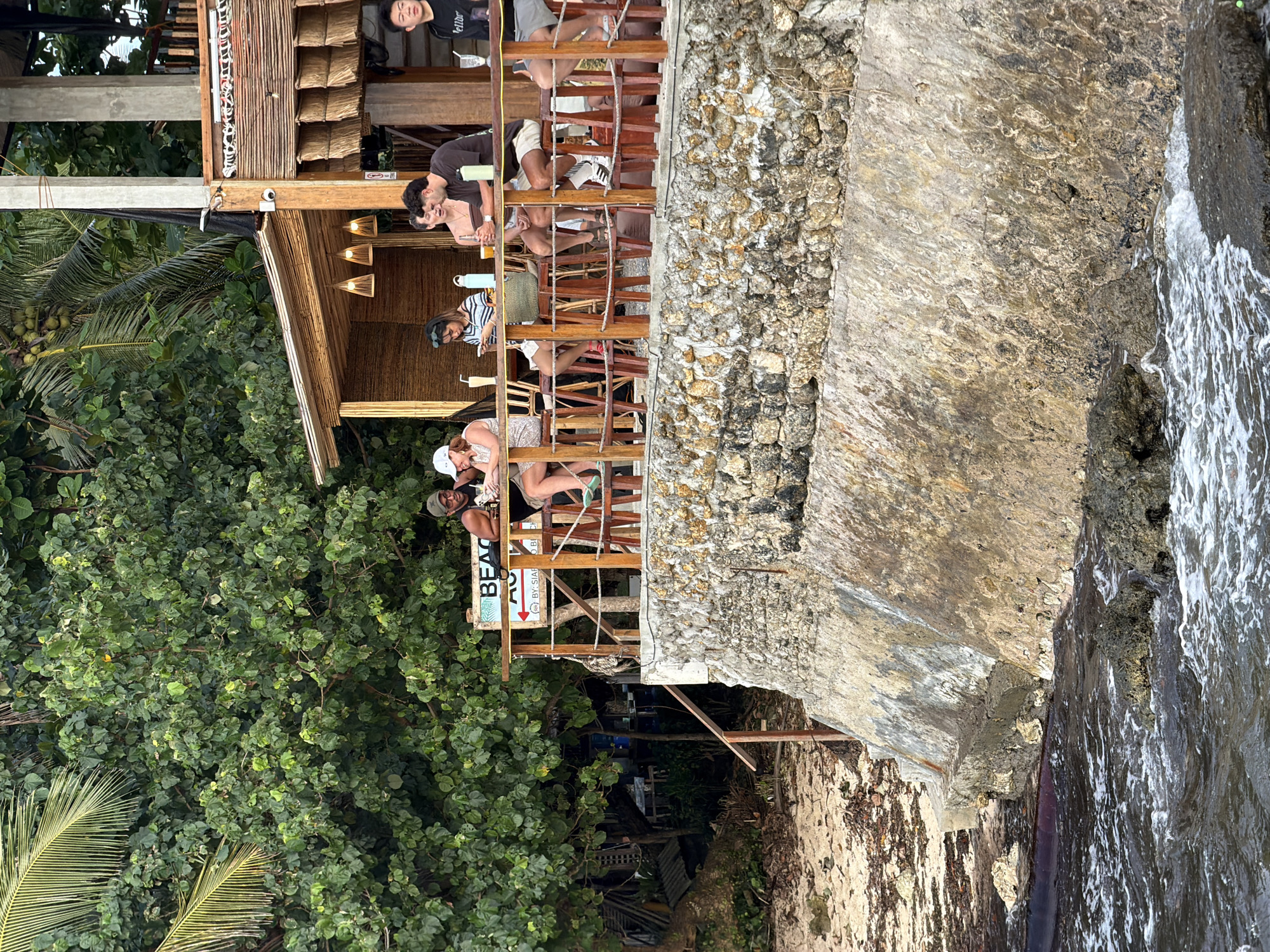 The image depicts a seaside bar/restaurant in General Luna, Philippines, built on a rocky outcrop over the ocean. The structure is multi-tiered with a thatched roof. Patrons are seated at the edge, interacting and dining. The foreground consists of dense tropical vegetation with a view of the ocean. A "Beach" sign is visible near the bottom of the image. A rocky wall is on the right side of the structure, with ocean waves splashing against it.