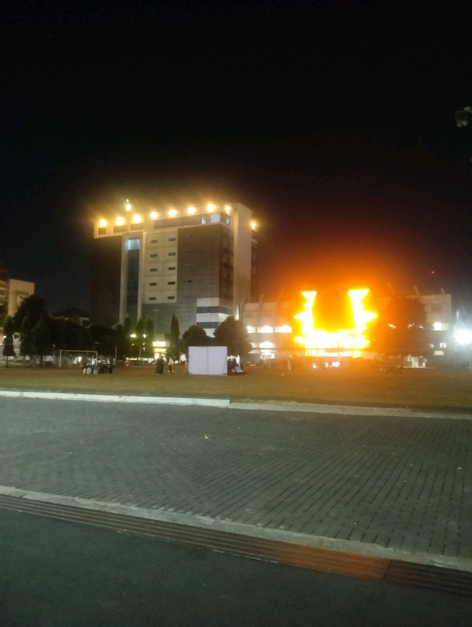 An outdoor scene at night in the city of Purwokerto, Indonesia, features a tall, multi-story building with illuminated windows and a prominent, brightly lit upper section. Adjacent to this building, a structure is intensely lit with a large, orange, U-shaped light display. In the foreground, a paved area with a distinctive pattern of dark gray interlocking blocks leads to a grassy field. On the field, several people are visible, along with what appear to be portable white structures and some vehicles, possibly golf carts. Trees are scattered across the field, and a soccer goal is visible in the background. The overall atmosphere suggests an event or gathering taking place at night.