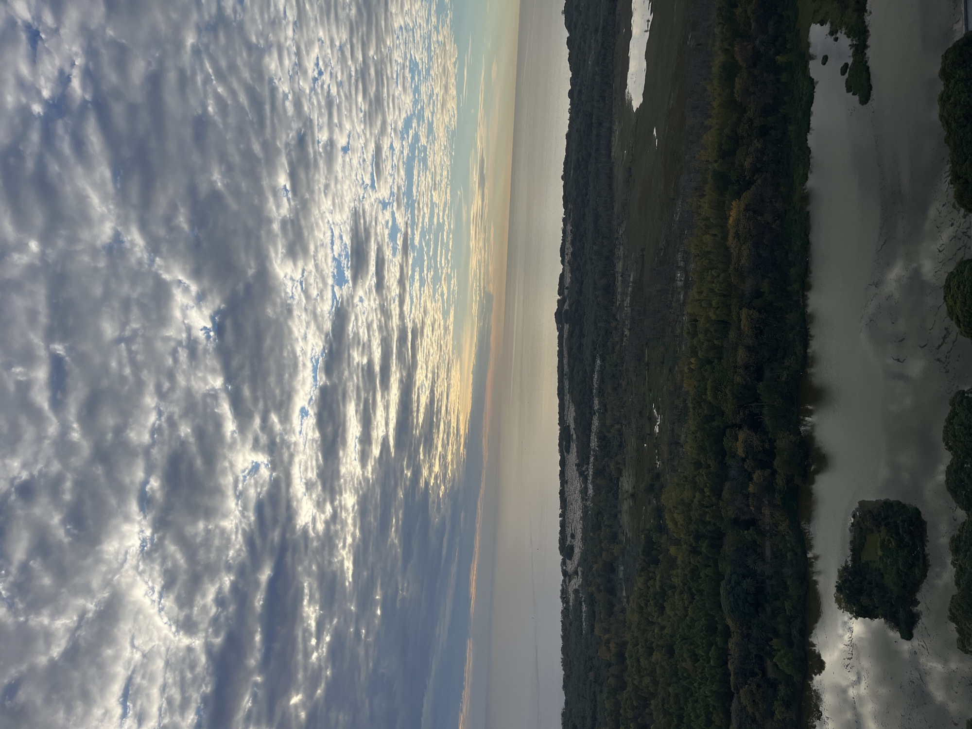 The image shows an aerial view of a landscape. On the left, there is a sky filled with rippled clouds illuminated by the sun. The central portion of the image depicts a body of water or river, possibly reflecting the sky. The right side of the image reveals a forested area with varied green tones and scattered water bodies or wetlands. There are no people visible in the image. The location information for the city of Buenos Aires, Argentina is not discernible from the image.
