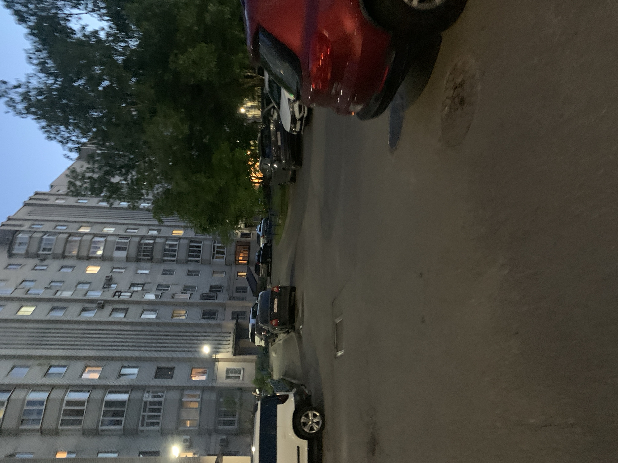 The image depicts a quiet urban residential street scene at dusk or early night, likely in Stavropol, Russia. On the left, a tall, multi-story apartment building with a distinctive grey facade and numerous rectangular windows dominates the frame. Its architecture appears characteristic of Soviet or post-Soviet era design, with many windows showing illuminated interiors, indicating occupancy within the building.
A paved asphalt street runs parallel to the building on the right, lined with various parked vehicles. In the immediate foreground, the rear quarter panel and wheel of a glossy red car are prominently visible. Further down the street, several other cars are parked bumper-to-bumper, including a white van with a blue stripe and multiple sedans in shades of black and grey. A black sedan in the mid-ground displays a partially visible Russian license plate, including characters such as 'В963'.
Lush green trees with dense foliage are scattered between the street and the apartment building, their tops silhouetted against the light blue-grey evening sky. The ambient light of twilight combines with artificial illumination from streetlights and the building's windows, casting a gentle glow over the scene. No people are visible, and the overall atmosphere is still and peaceful. The street surface appears largely clear, with some texture from the asphalt and a circular utility cover visible near the red car.