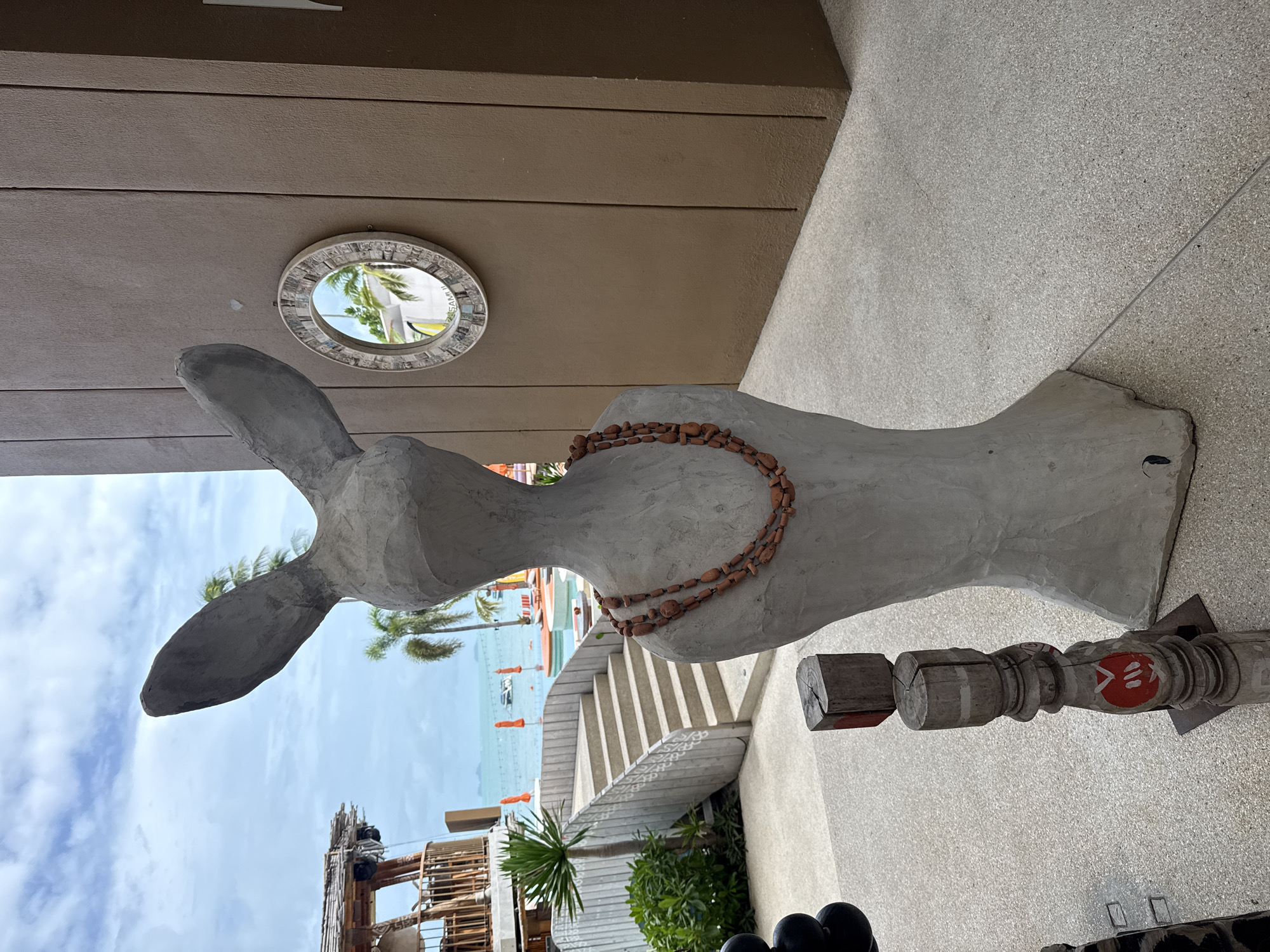 A grey, humanoid sculpture with large rabbit-like ears stands in the foreground, wearing a necklace of brown beads. To the left of the sculpture, a wooden post with carved decorative elements and red markings stands on the speckled grey pavement. In the background, a modern building with a textured brown wall is visible, featuring a circular mirror with a decorative frame. Beyond that, a swimming pool with orange umbrellas and lounge chairs is set against a backdrop of palm trees and a body of water, with a clear blue sky overhead. The overall setting appears to be a resort or outdoor dining area in Ko Samui District, Thailand, likely near the coast.
