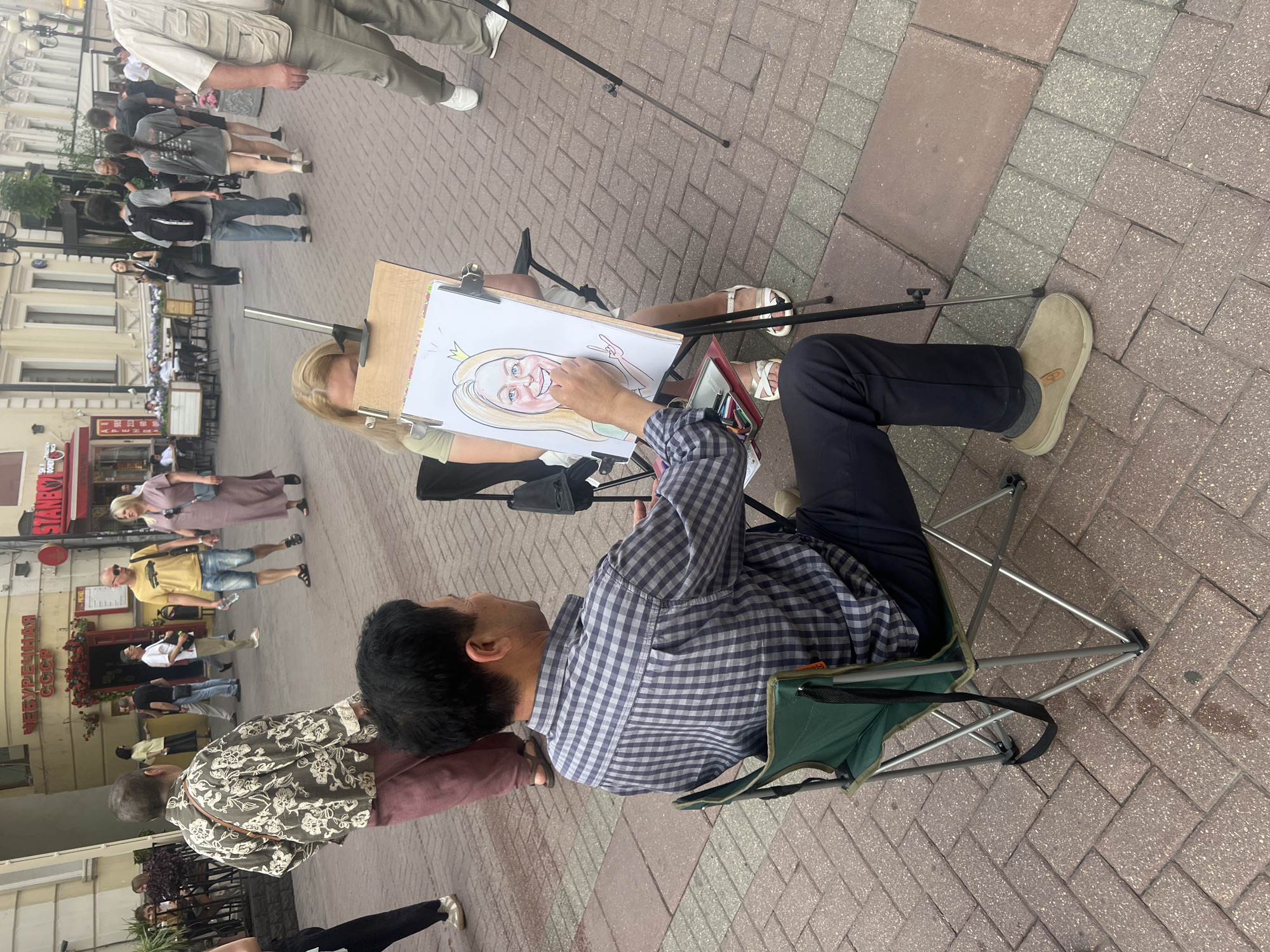 The image depicts a street scene in Moscow, Russia. In the foreground, an Asian man is seated in a folding green chair, drawing a caricature of a blonde woman who is sitting in front of him, facing away from the camera. The drawing shows a cartoonish portrait of the woman with exaggerated features, and the man is in the process of adding detail with a pencil. The woman is wearing a light green top and white sandals. The man is wearing a blue and grey checkered shirt and dark pants.
In the background, pedestrians are walking along a brick-paved street lined with buildings. One building has Cyrillic signage that reads "ЧЕБУРЕЧНАЯ СССР" (Cheburechnaya USSR). Other signs and architectural details suggest an urban commercial area. People in the background are dressed in casual summer clothing. The overall atmosphere is that of a lively outdoor marketplace or pedestrian zone.