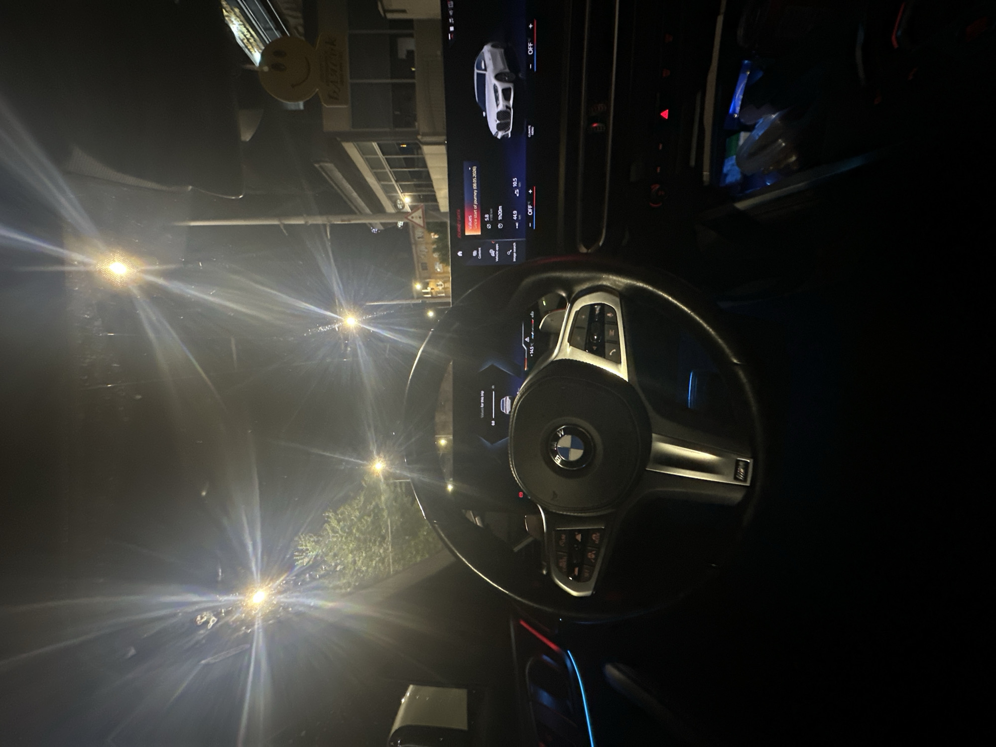 Here is a matter-of-fact description of the scene:
The image depicts the interior of a BMW car at night. The steering wheel, with the BMW logo visible, is the central focus. Behind the steering wheel, the car's dashboard displays information such as "Values" and journey details. Outside the car window, street lights illuminate what appears to be an urban environment. A smiley face decoration hangs from the rearview mirror.