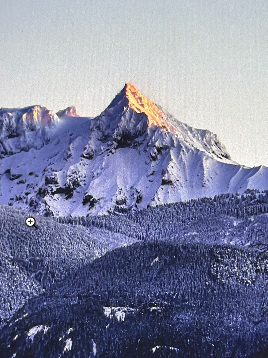 The image displays a mountainous landscape covered in snow. The peak of the tallest mountain in the center is illuminated by the golden light of the sun, creating a striking contrast with the blue-tinged snow on the surrounding slopes. In the foreground, a dense forest of snow-covered trees blankets the lower parts of the mountains. A small, circular zoom icon with a plus sign is visible on the left side of the image, overlaid on the forested area, suggesting a digital interface element. The sky above is a pale, washed-out blue, indicating either early morning or late afternoon. The image appears to be a photograph taken of a computer screen, as evidenced by the slight graininess and the presence of the zoom icon. There is no indication of the city of Kafr Yasif, Israel, in this image; it depicts a natural, alpine environment.