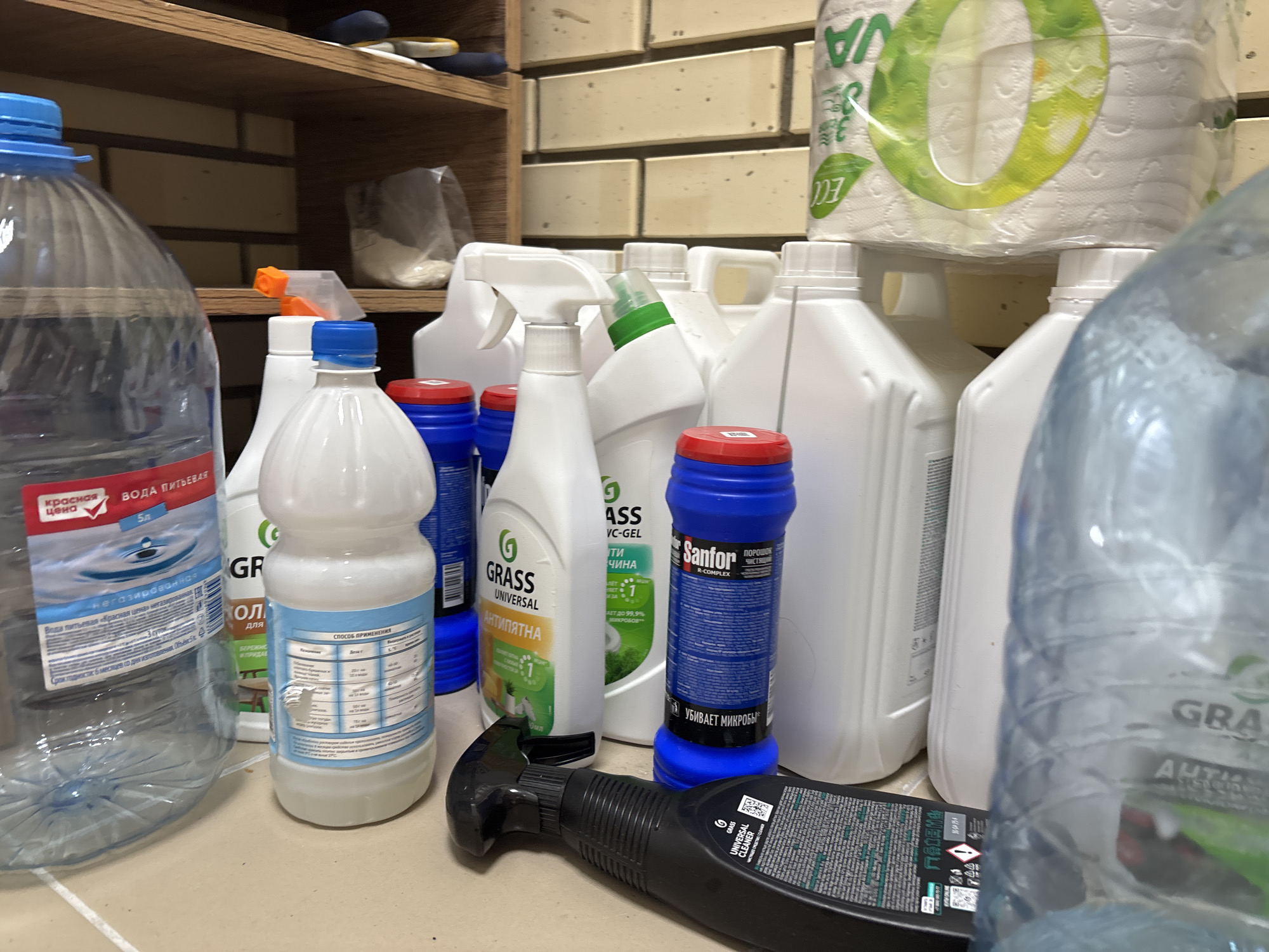 The scene is an indoor utility or storage area in Karasi, Russia, featuring an assortment of household cleaning products and water bottles. These items are arranged on a light-colored, smooth surface, with a brick-patterned wall and wooden shelving in the background.
On the far left, a transparent 5-liter plastic bottle with a blue screw cap is visible. Its red and white label displays "КРАСНАЯ ЦЕНА" (Red Price) and "ВОДА ПИТЬЕВАЯ 5Л" (Drinking Water 5L). To its right, an opaque white plastic bottle with a blue cap and a light blue label shows partial text including "СПОСОБ ПРИМЕНЕНИЯ" (Method of Application) in a table format.
A black plastic trigger spray bottle, labeled "GRASS UNIVERSAL CLEANER" and featuring a QR code, lies horizontally in the foreground. Behind it, a cluster of various white plastic bottles, likely containing cleaning solutions, are visible. One prominent white bottle has a green push-pull cap and a green label with the "GRASS" logo and "UNIVERSAL АНТИПЯТНА" (ANTI-STAIN) text. Another white bottle is fitted with a white trigger spray head.
A blue cylindrical container with a red cap is present among the white bottles. Its label identifies it as "SANFOR R-COMPLEX ПОРОШОК ЧИСТЯЩИЙ" (Cleaning Powder) and states "УБИВАЕТ МИКРОБЫ" (KILLS MICROBES). Resting on top of two of the white cleaning bottles is a single roll of white toilet paper, encased in clear plastic packaging that displays green and white branding, including text suggestive of "ECO" and an indication of "3 layers". A partially visible, out-of-focus clear plastic bottle, similar to the 5L water bottle, is on the far right.
The background consists of a wall constructed from light-colored, horizontal rectangular bricks or tiles. Above this wall, dark wooden shelves are present, with indistinct items visible on them. No people are present, and no actions are occurring in the static scene.