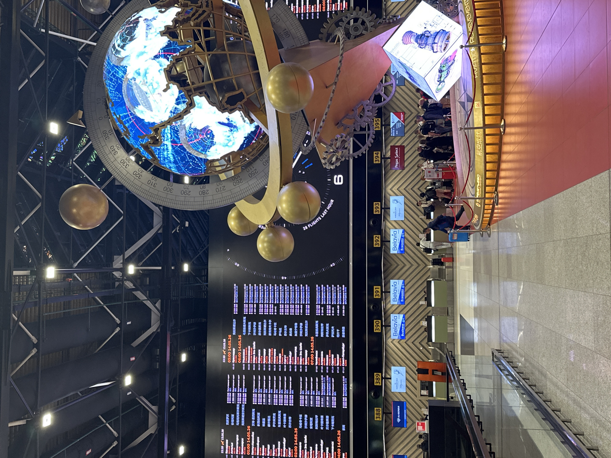 The image depicts a scene inside what appears to be an airport in Khimki, Russia. Overhead is a large, decorative structure with an illuminated globe, golden spheres, and mechanical gears. Below, a large flight information display shows numerous flight details in Russian and possibly Chinese. In the lower right of the image is a section indicating "Aviation Achievements" and a queue of people. Flight check-in counters labeled 388 through 395 and displaying airline names are visible. The counters display airline logos.