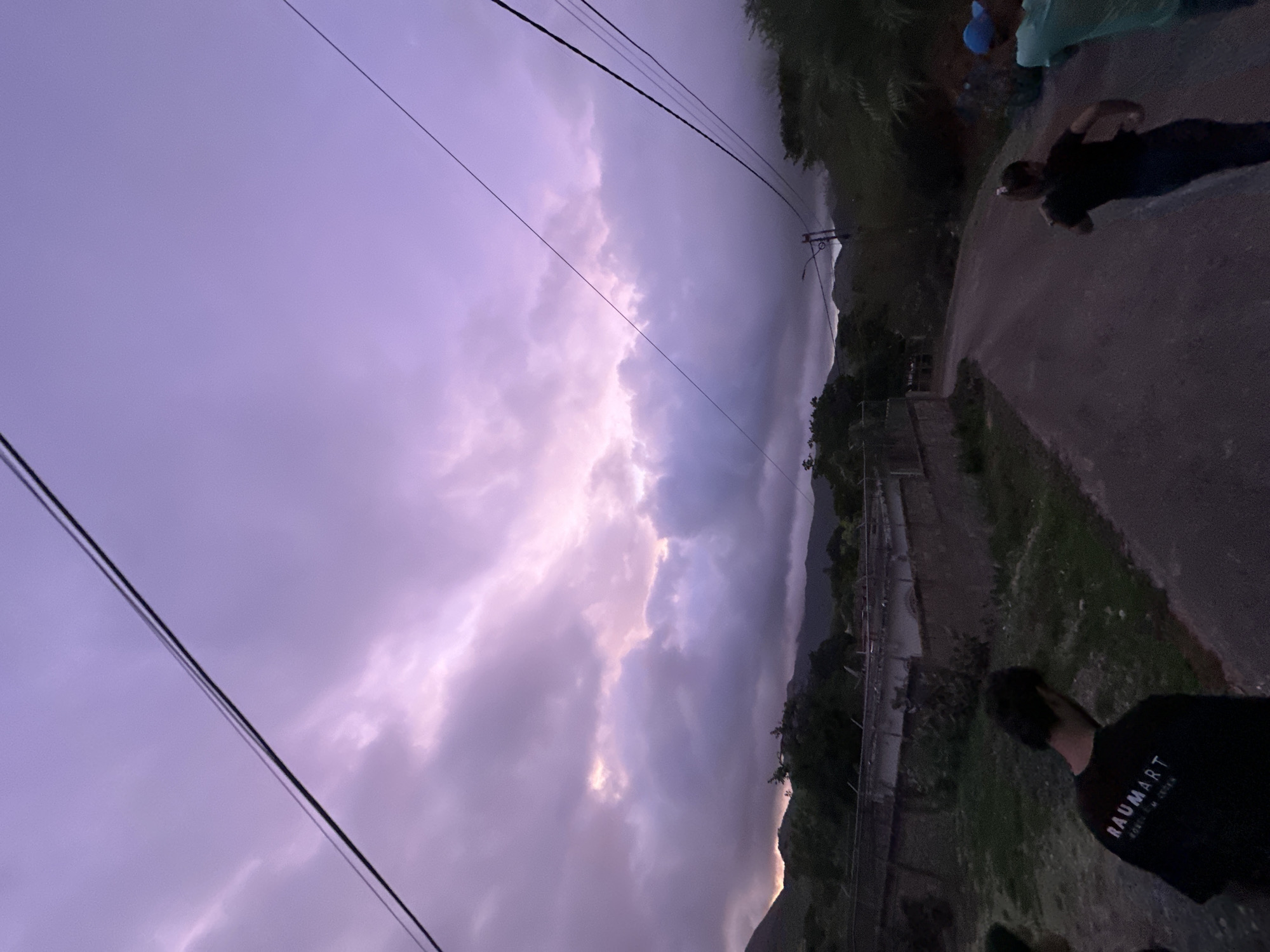 The image depicts an outdoor scene in La Asuncion, Venezuela, at dusk. The sky is a dramatic mix of purple and pink, with heavy clouds suggesting an impending storm or the aftermath of rain. Power lines traverse the upper portion of the frame, casting faint shadows. In the foreground, a paved road curves gently into the distance, flanked by vegetation and a low concrete structure on the left. Several individuals are visible. On the left side, a person wearing a black t-shirt with the text "RAUMART" is partially visible from the back. Further down the road, two figures are running away from the camera; one is wearing a dark shirt and dark pants, and the other is wearing a light blue shirt and a blue cap. The surrounding environment includes hilly terrain covered in trees in the background. The overall atmosphere is subdued due to the low light conditions characteristic of twilight.